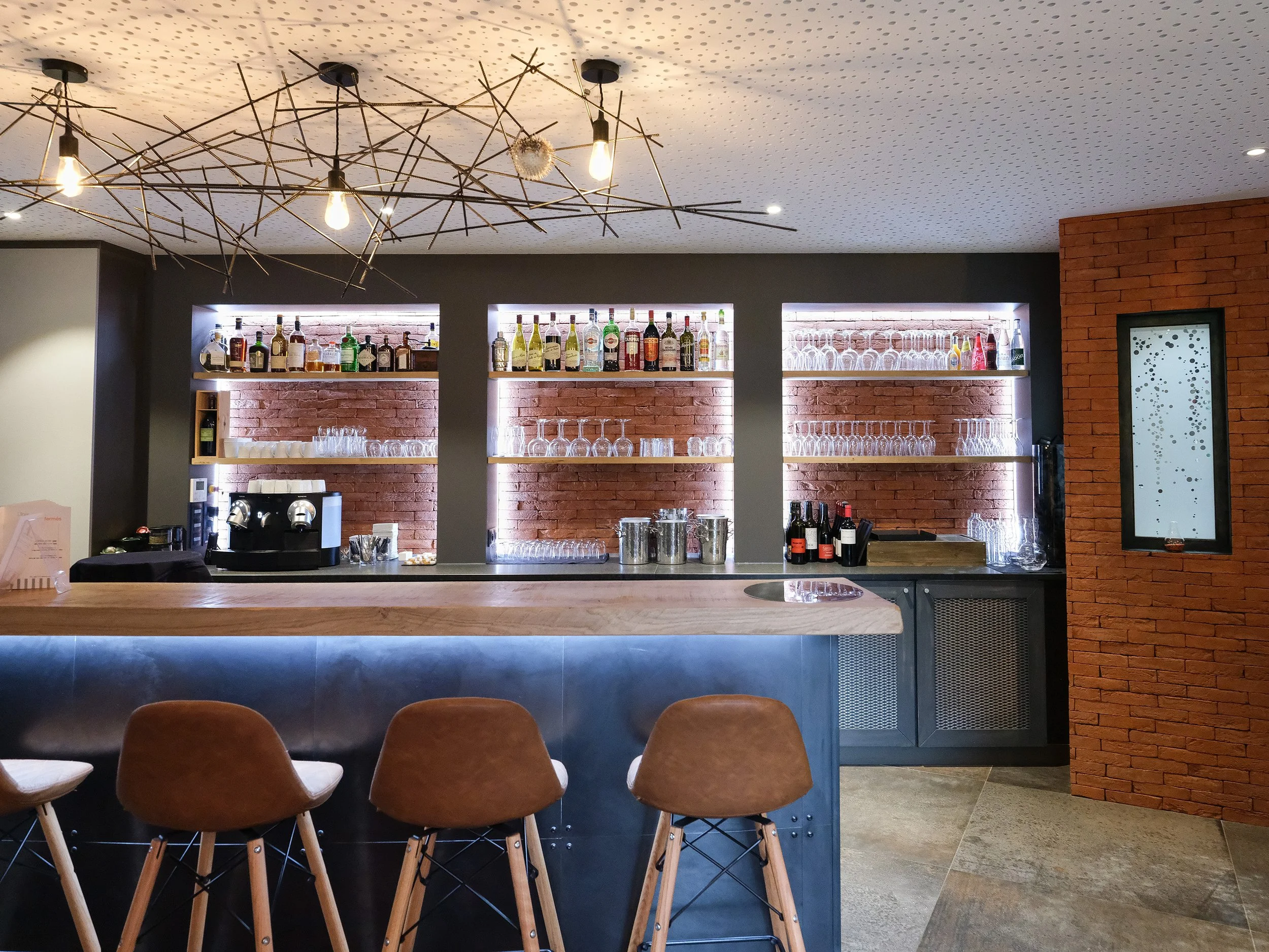 Bar moderne avec une rangée de chaises en face, étagères en brique avec bouteilles d'alcool et verres rétroéclairés, plafond avec lumière décorative.