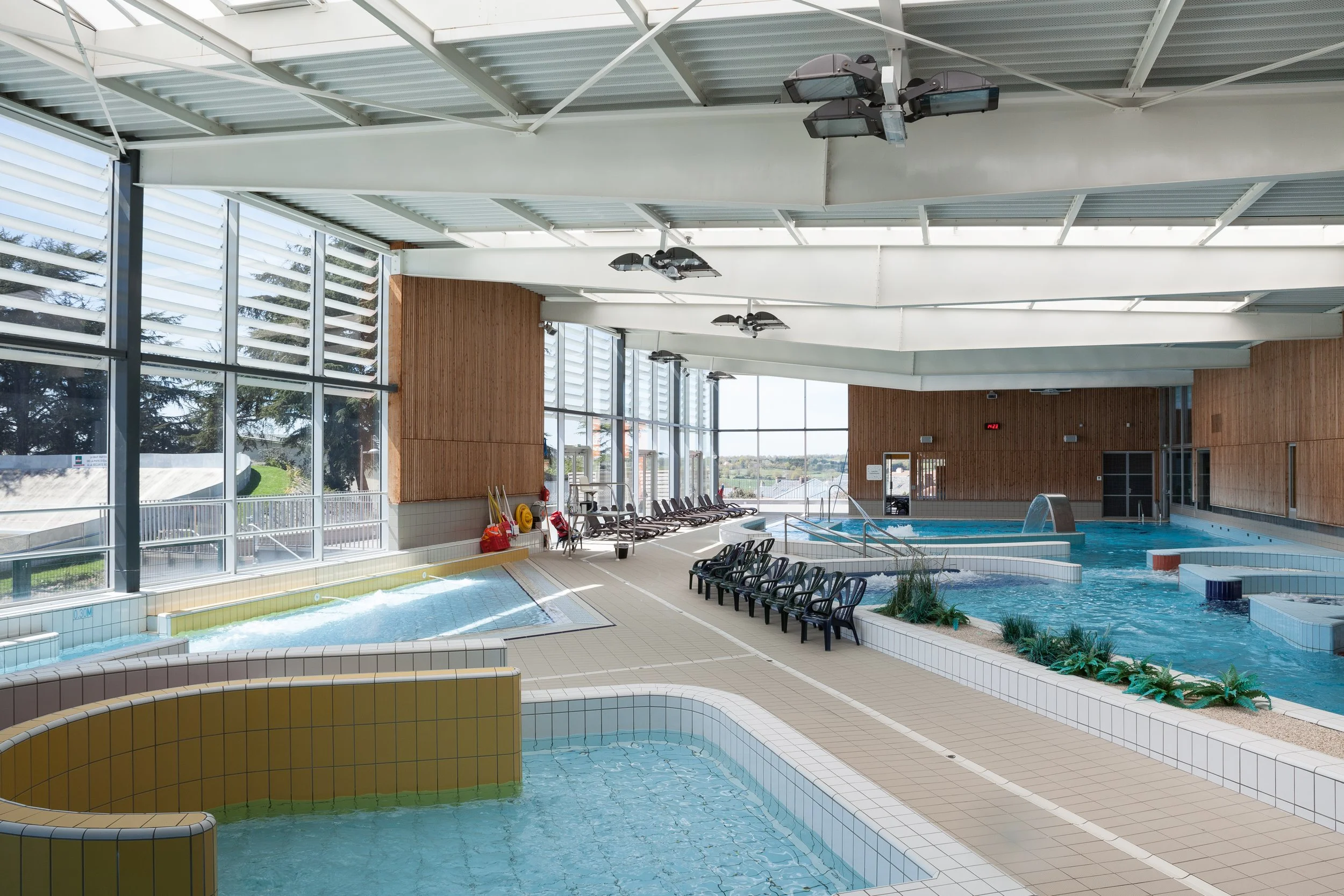 Piscine intérieure avec plusieurs bassins, chaises longues, et fenêtres panoramiques offrant la vue sur l'extérieur, éclairée par la lumière naturelle.