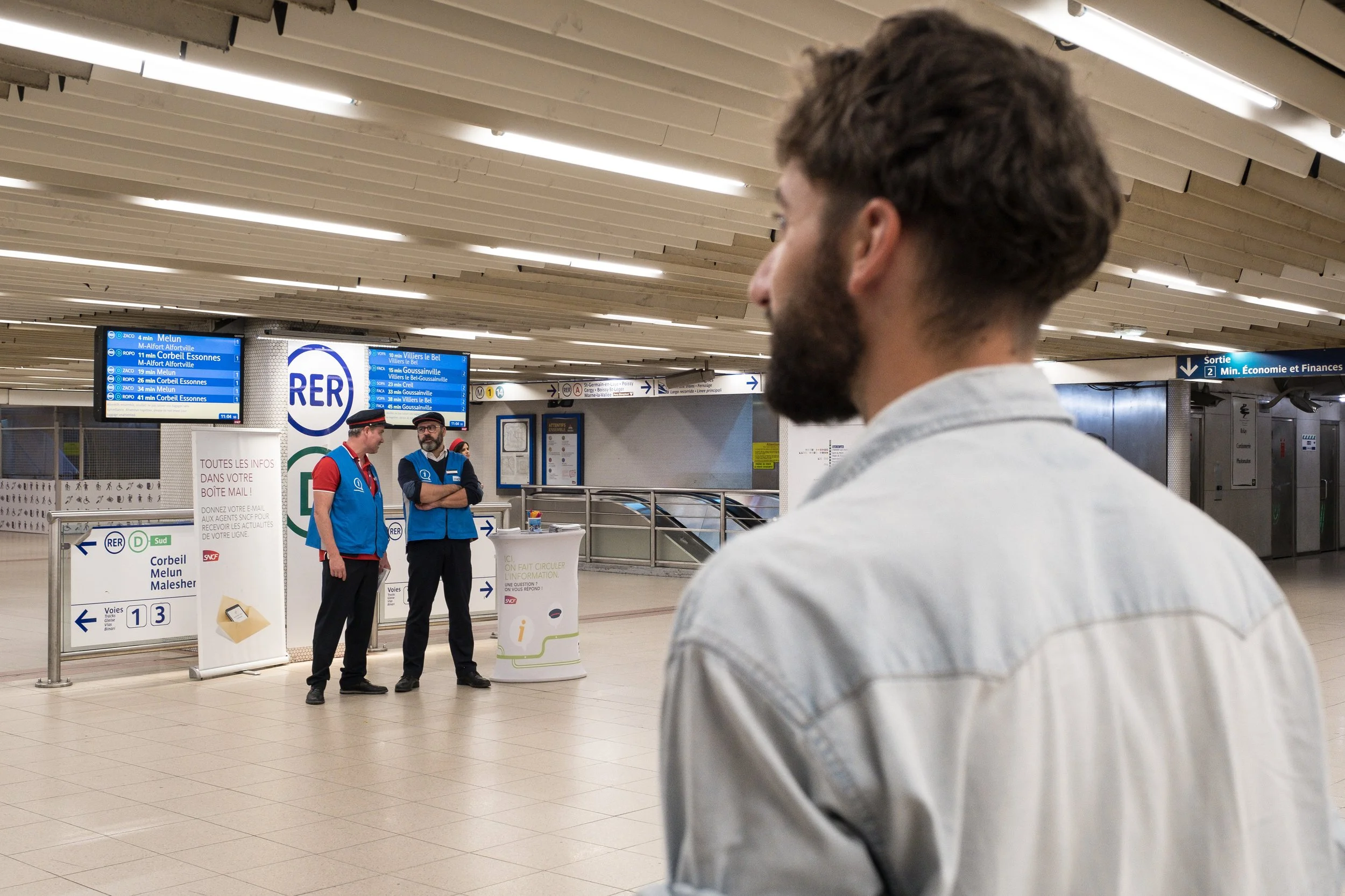 Un homme avec une barbe et une chemise claire dans une gare, regardant vers le groupe de deux personnes en vestes bleues discutant à l'arrière-plan.