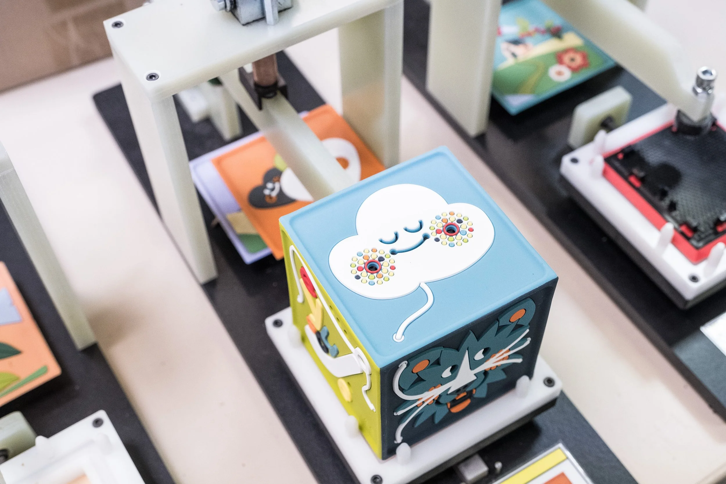 Un cube en plastique coloré avec une face représentant un visage de lion, décoré de motifs floraux et un visage souriant, placé sur une surface surmontée d'une structure mécanique.