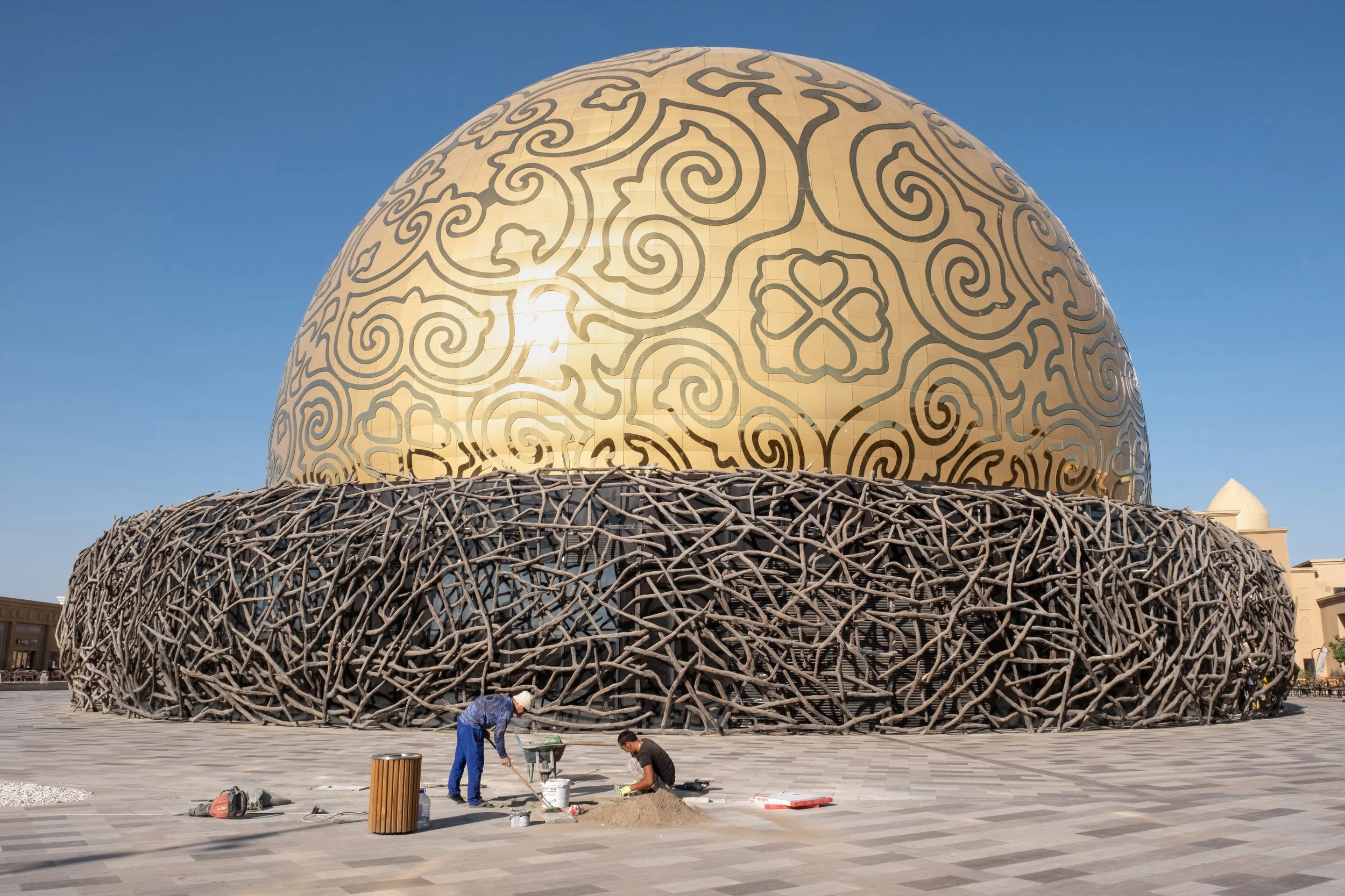 Deux ouvriers travaillent sur un sol pavé devant une grande structure en forme de dôme doré avec des motifs noires délicats, entourée d'une structure en bois ressemblant à un nid à Turkestan, au Kazakhstan.