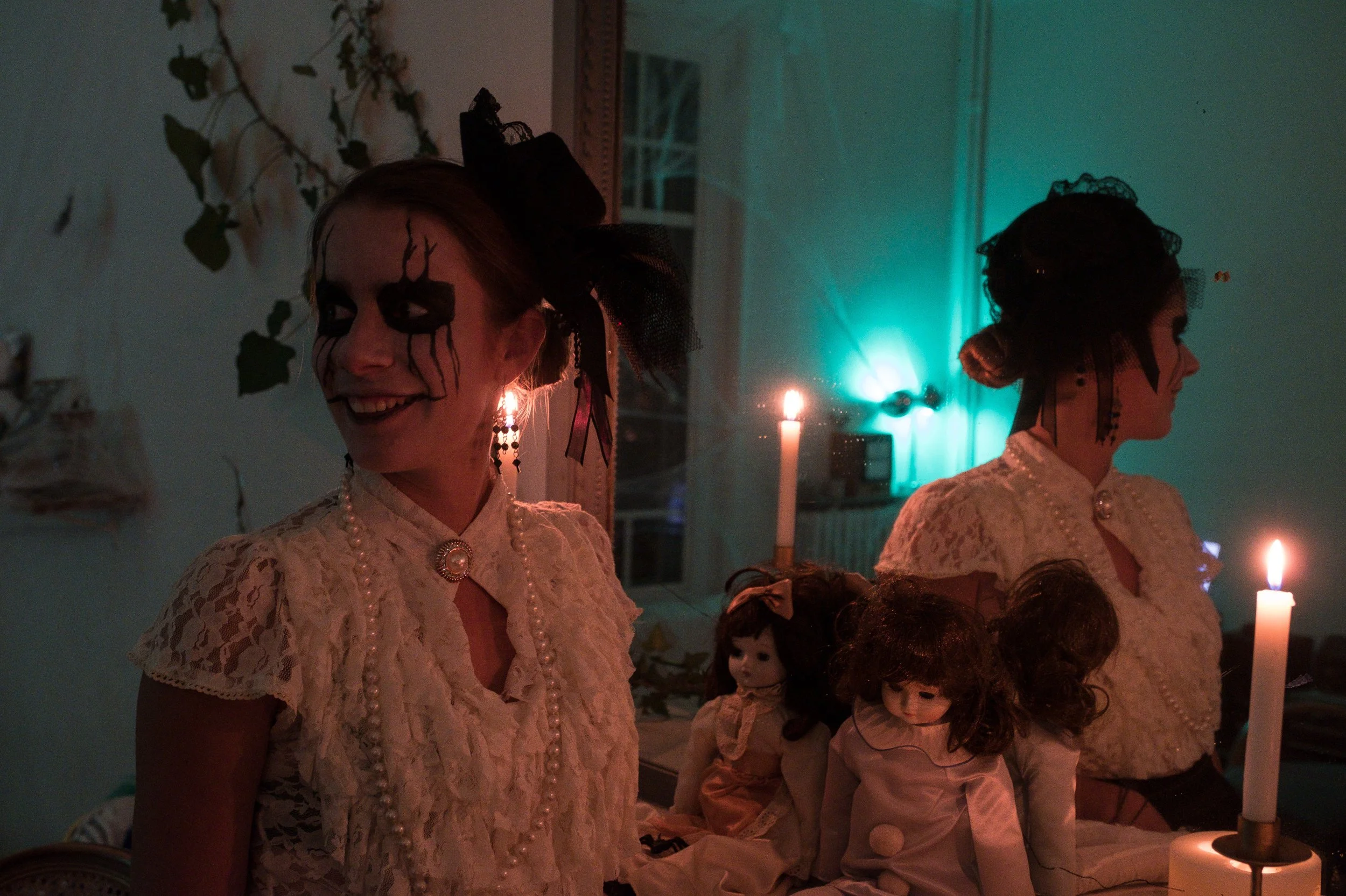 Une femme avec un maquillage effrayant et un costume de Halloween, portant une robe en dentelle blanche et un collier de perles, sourit dans une pièce sombre éclairée par des bougies et une lumière verte, entourée de poupées anciennes.