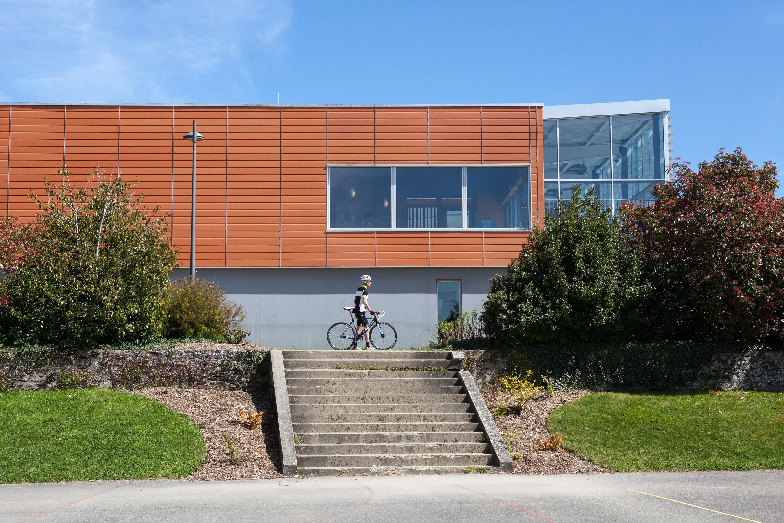 Un cycliste portant un casque et un gilet réfléchissant, montant une rampe d'escalier en béton devant un bâtiment moderne.