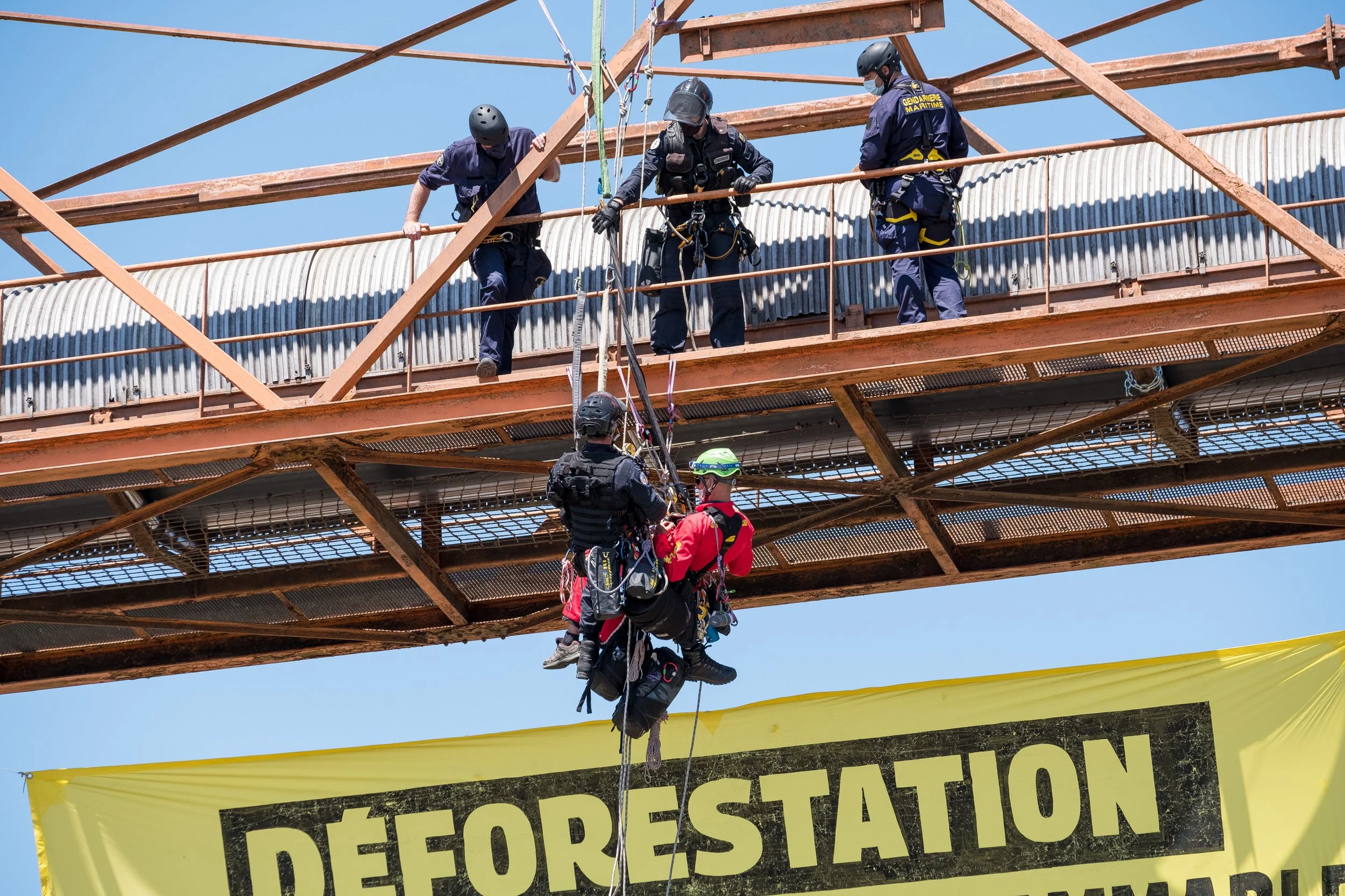 Des forces de l'ordre en train de réaliser une opération d'alpinisme pour arrêter des activistes de Greenpeace, sous un ciel clair, avec un panneau jaune portant le mot 'Déforestation' en français.