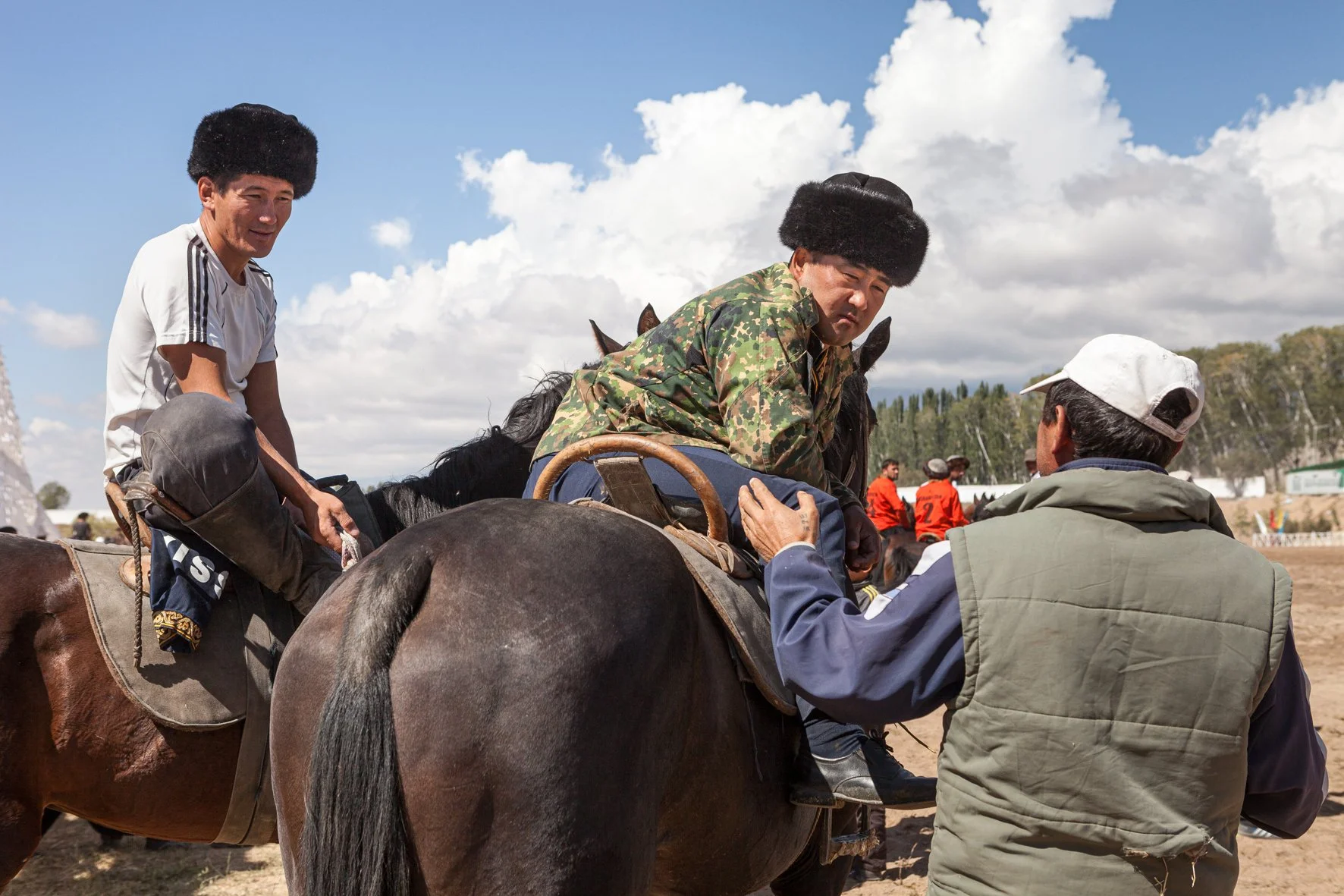 Deux hommes en tenue traditionnelle et un homme en blouson conversent près d'un cheval, avec un groupe de personnes en arrière-plan sous un ciel partiellement nuageux.