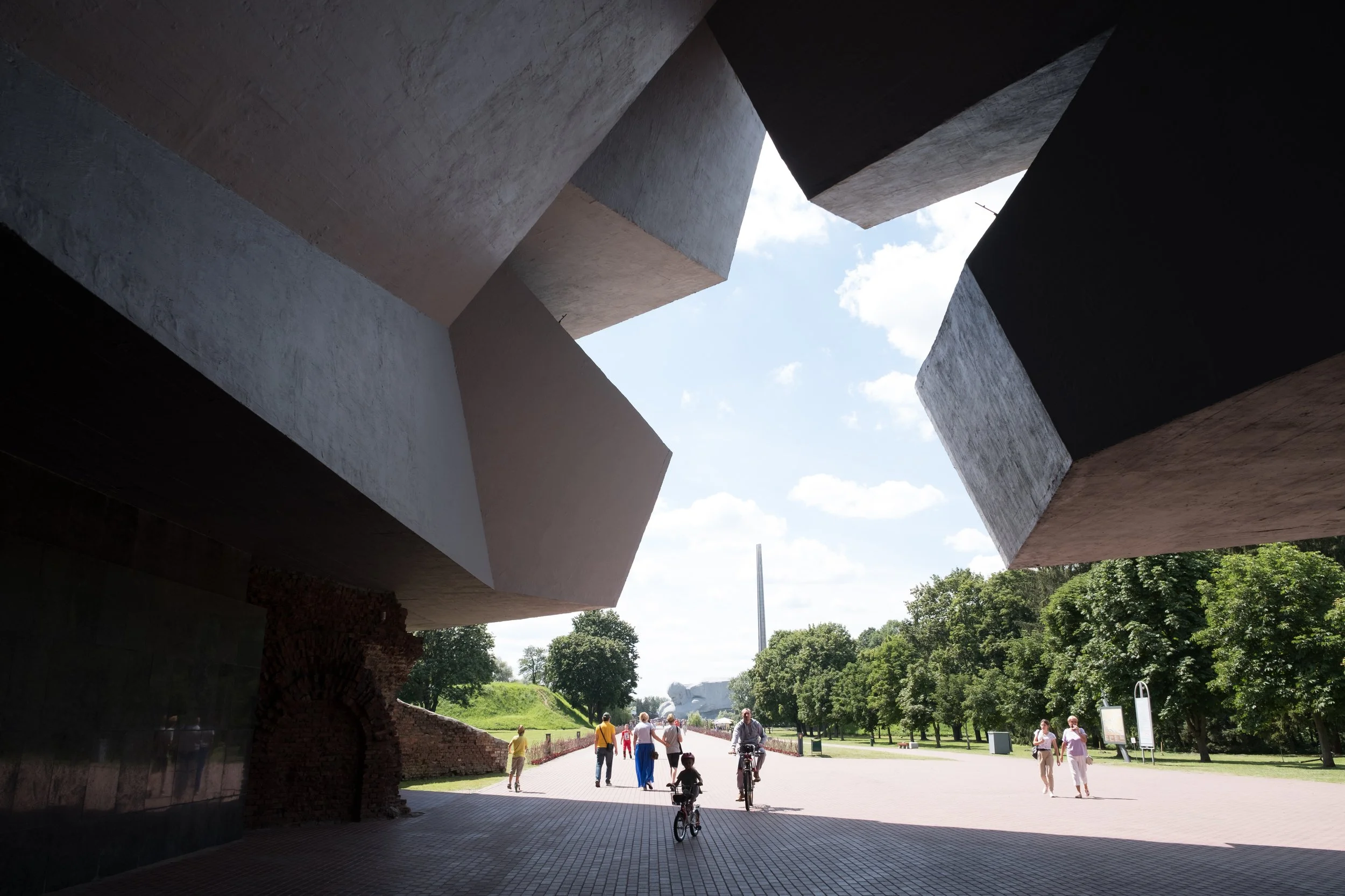 Vue en contre-plongée d'une structure architecturale moderne en béton avec des formes géométriques angulaires, ouverte vers un parc avec des personnes qui se promènent en Biélorussie.
