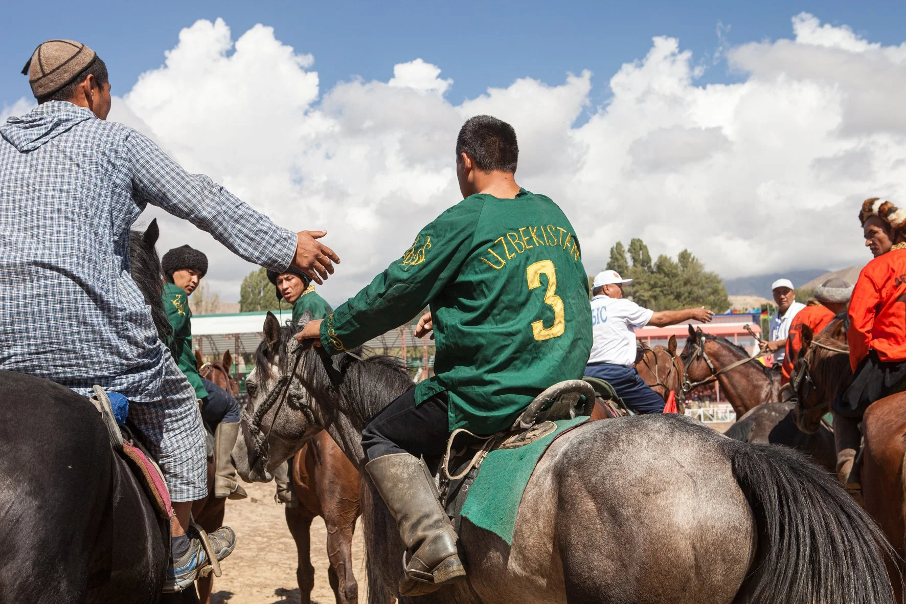 Groupe de cavaliers en tenue traditionnelle, certains portant des chapeaux, en plein match de kok boru dans un espace en plein air sous un ciel nuageux.