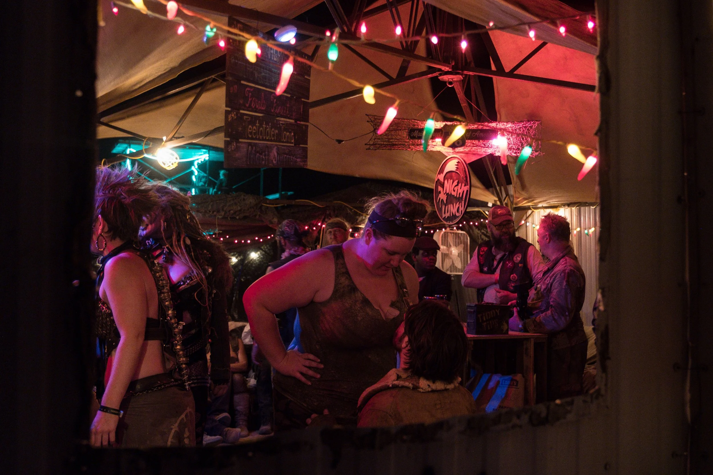 Une scène de bar avec des personnes buvant des shots depuis la poitrine d'une femme, entourés d'autres personnes dansant et discutant dans une tente éclairée de lumières colorées, ambiance chaleureuse et festive.