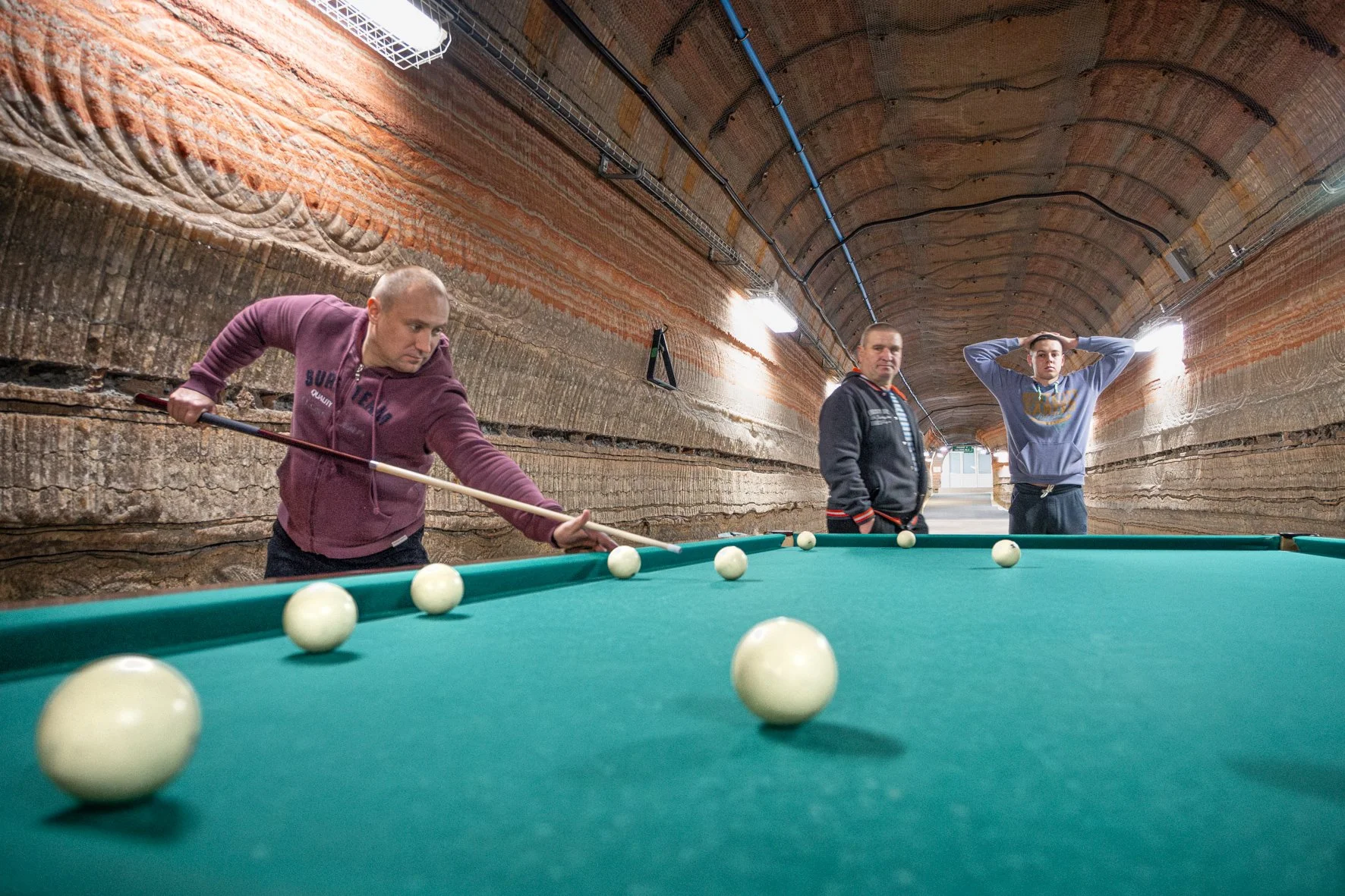 Trois hommes jouent au billard dans un tunnel avec murs en couches de roche rouge orangée.