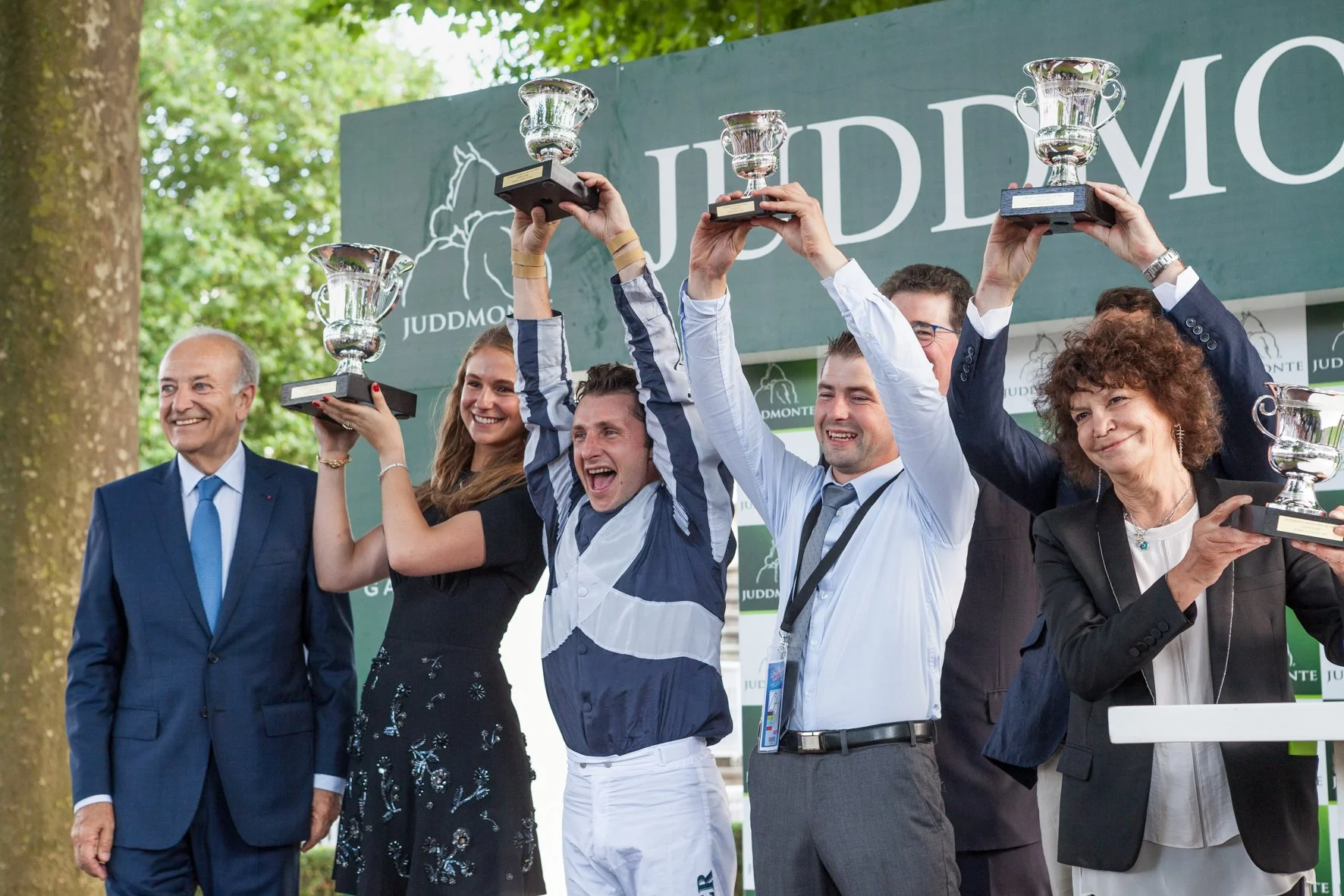 Groupe de personnes souriantes tenant des trophées lors d'une cérémonie de remise de prix en plein air avec un panneau 'JUDDMONT' en arrière-plan.