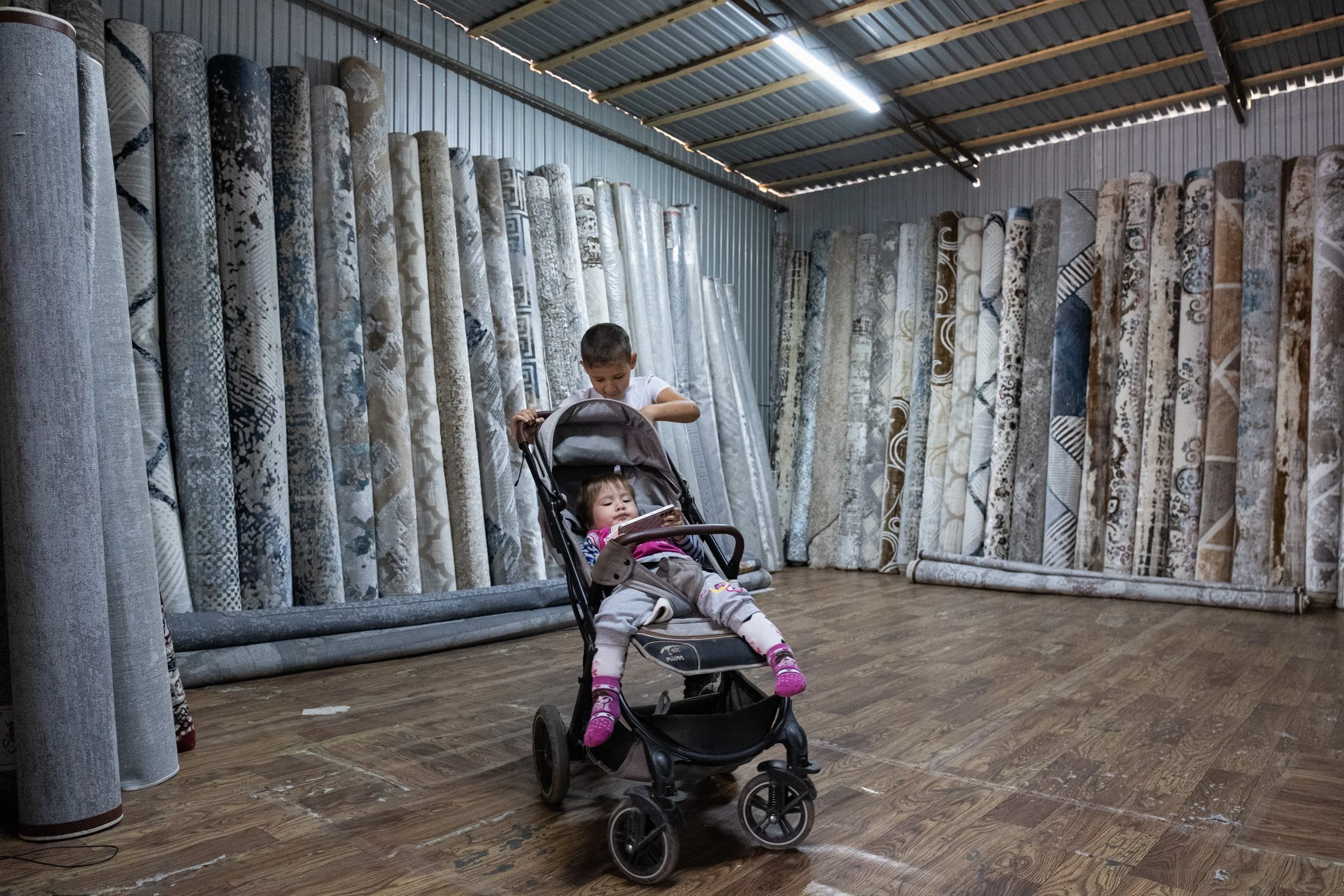 Deux enfants, un garçon et une petite fille, dans un magasin de tapis au Kazakhstan. La fille est assise dans une poussette et regarde un smartphone, le garçon fait rouler la poussette. Des rouleaux de tapis sont alignés au mur en arrière-plan.