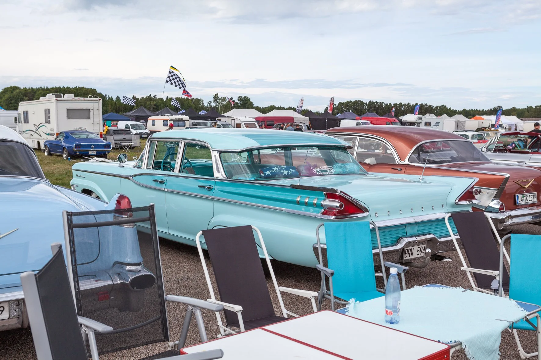 Voitures classiques en exposition lors d'un rassemblement auto, avec des chaises et une table de pique-nique dans le premier plan.