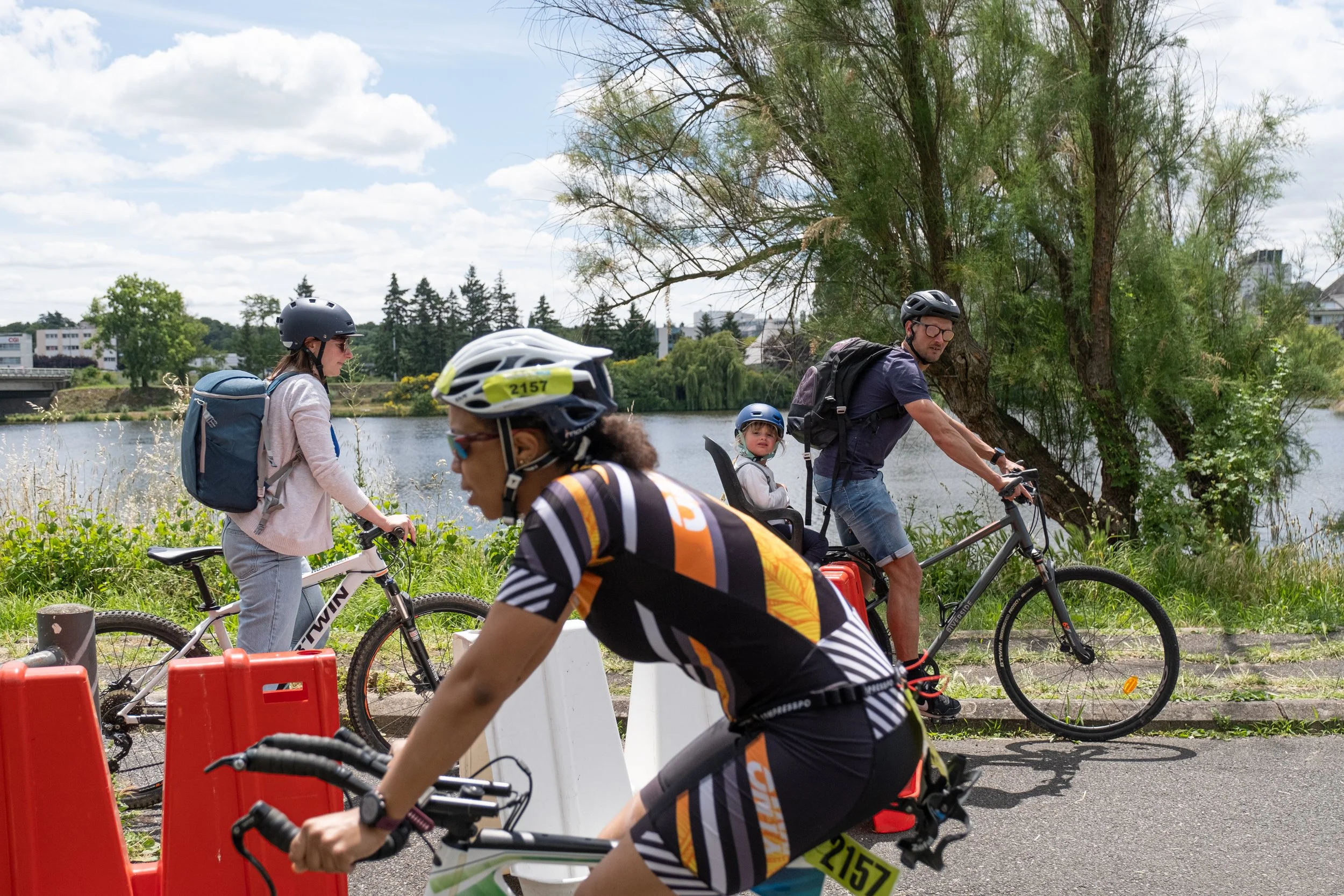 Groupe de personnes faisant du vélo près d'une rivière, avec des arbres et un ciel nuageux en arrière-plan.
