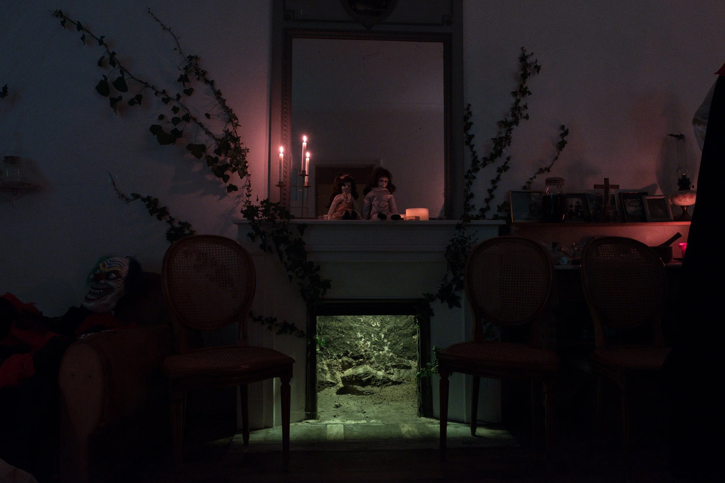 Une pièce sombre avec un miroir reflétant deux poupées posées sur une étagère, des bougies allumées. Une ouverture en bas au centre montre un chemin de pierres entouré de végétation. Des chaises sont disposées devant la cheminée, et une tête de clown