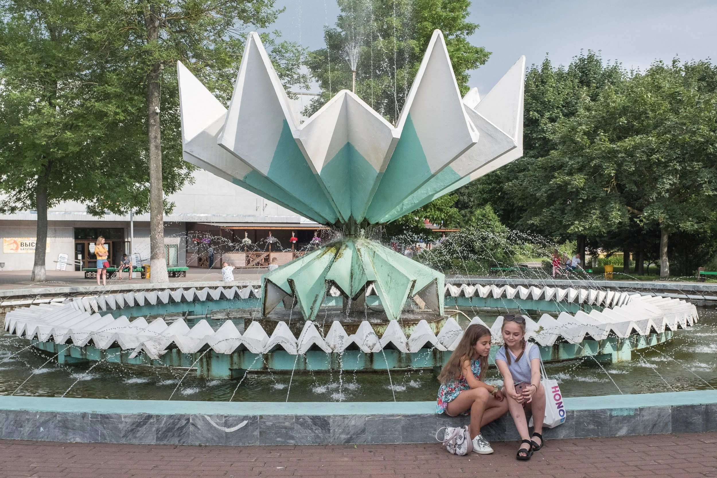 Deux jeunes filles assises au bord d'une fontaine avec une sculpture soviétique de lotus stylisé au centre, entourée d'eau, dans un parc avec des arbres et d'autres personnes en arrière-plan en Biélorussie.