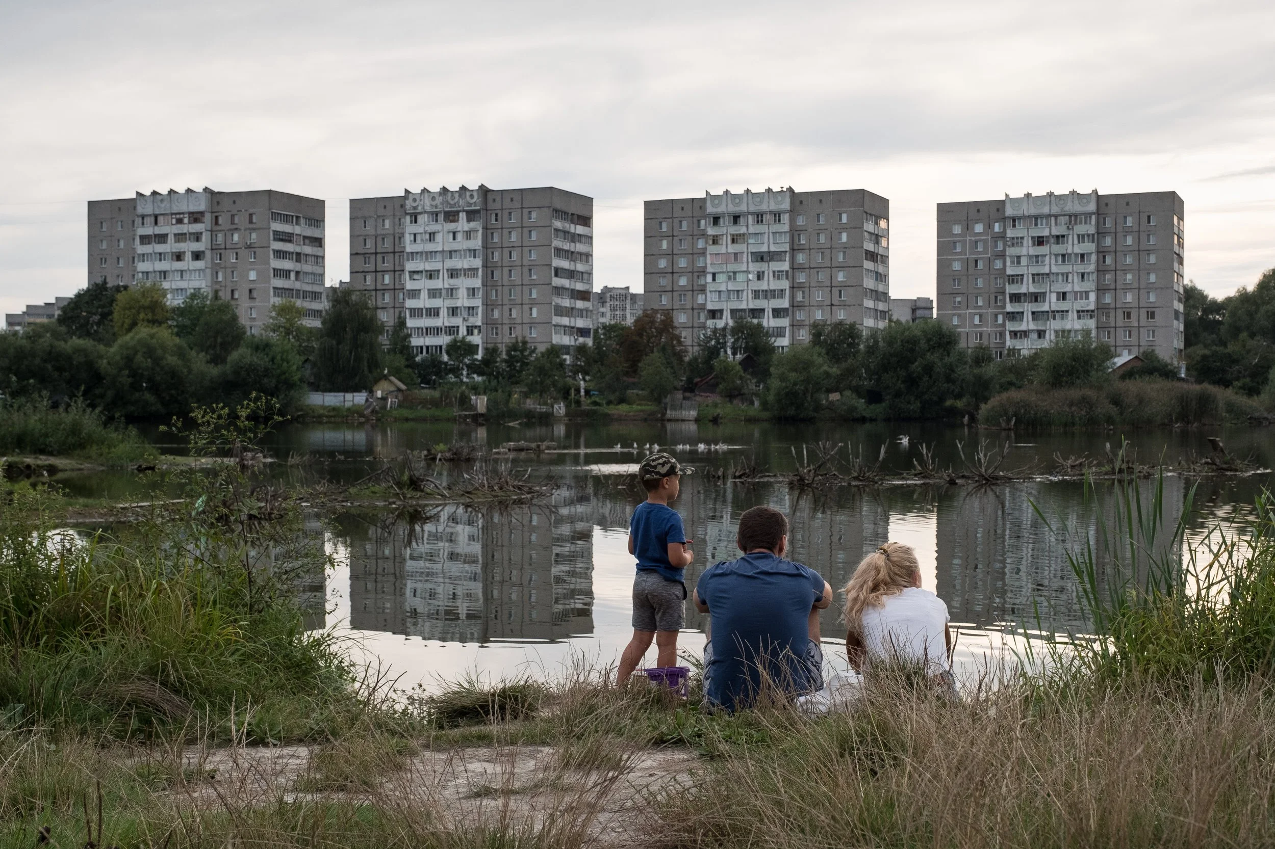 Un groupe de trois personnes, deux adultes et un enfant, au bord d'un lac, avec des immeubles résidentiels en arrière-plan, au coucher du soleil en Biélorussie.