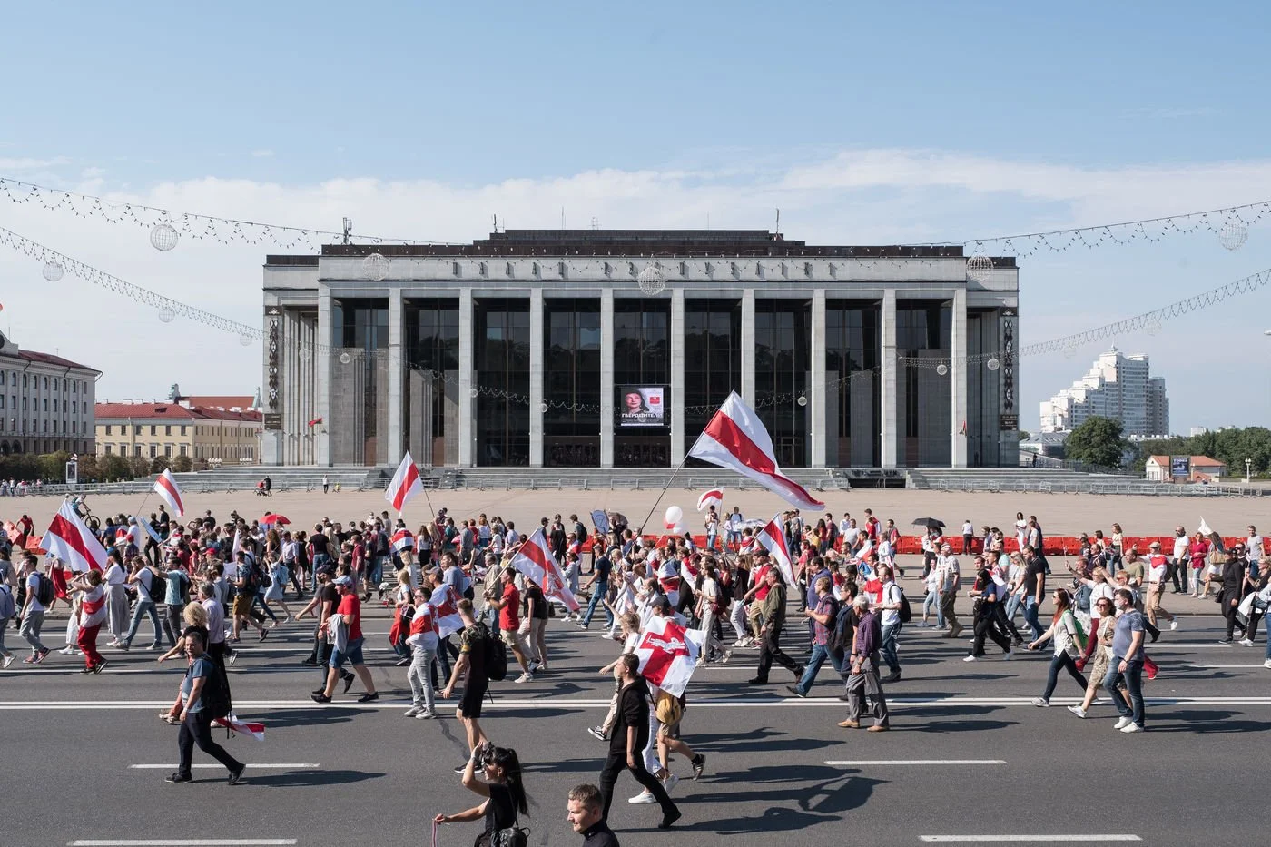 Foule marchant avec des drapeaux à rayures horizontales rouges et blanches devant un grand bâtiment moderne en pierre, avec des guirlandes décoratives suspendues dans le ciel clair.