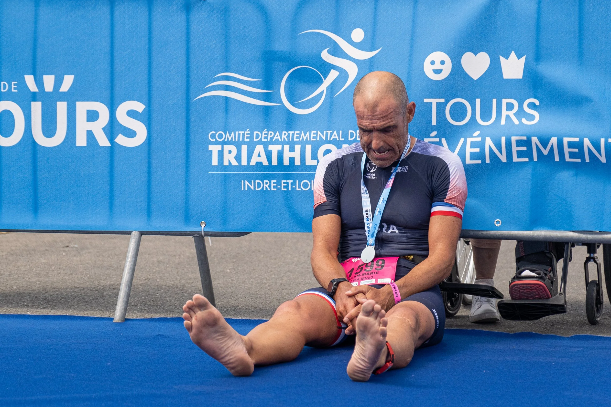 Un homme enkimé, assis par terre après un triathlon, portant une médaille, un dossard, et des vêtements sportifs, en train de souffrir ou d'être épuisé, devant une bannière bleue avec du texte en français.