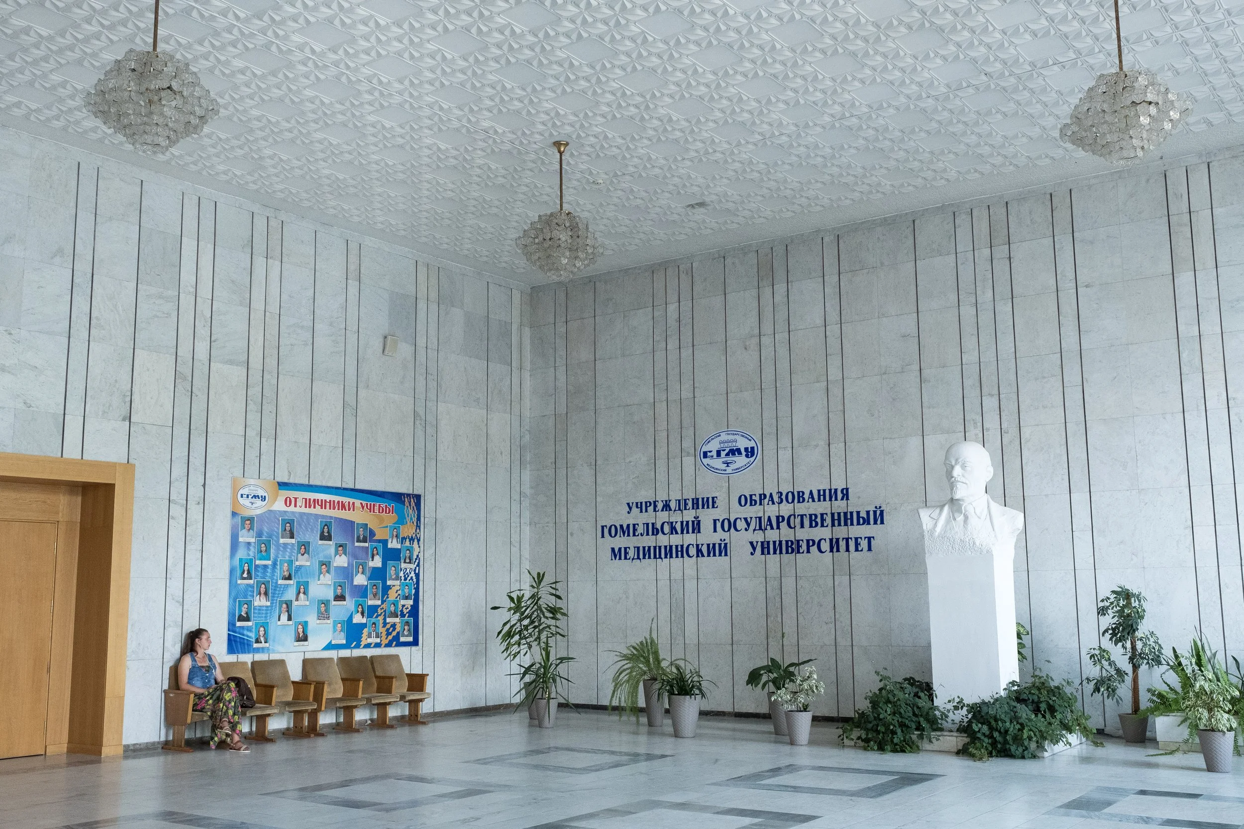 Hall d'entrée de l'université d'État de médecine de Gomel avec un mur en marbre, un buste blanc d'une statue d'homme, des plantes en pot, un tableau d'affichage avec des photos d'étudiants, et une femme assise sur une banquette.