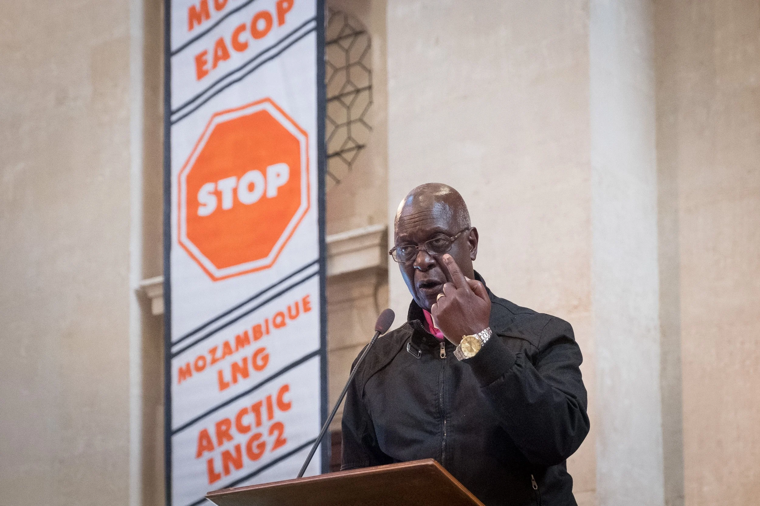 Un homme noir porte des lunettes et un bracelet en or, il se tient derrière un pupitre, faisant un geste avec sa main. En arrière-plan, un panneau affiche un symbole d'arrêt avec le texte "STOP" et des mots en portugais tels que "EACOP", "MOZAMBIQUE 