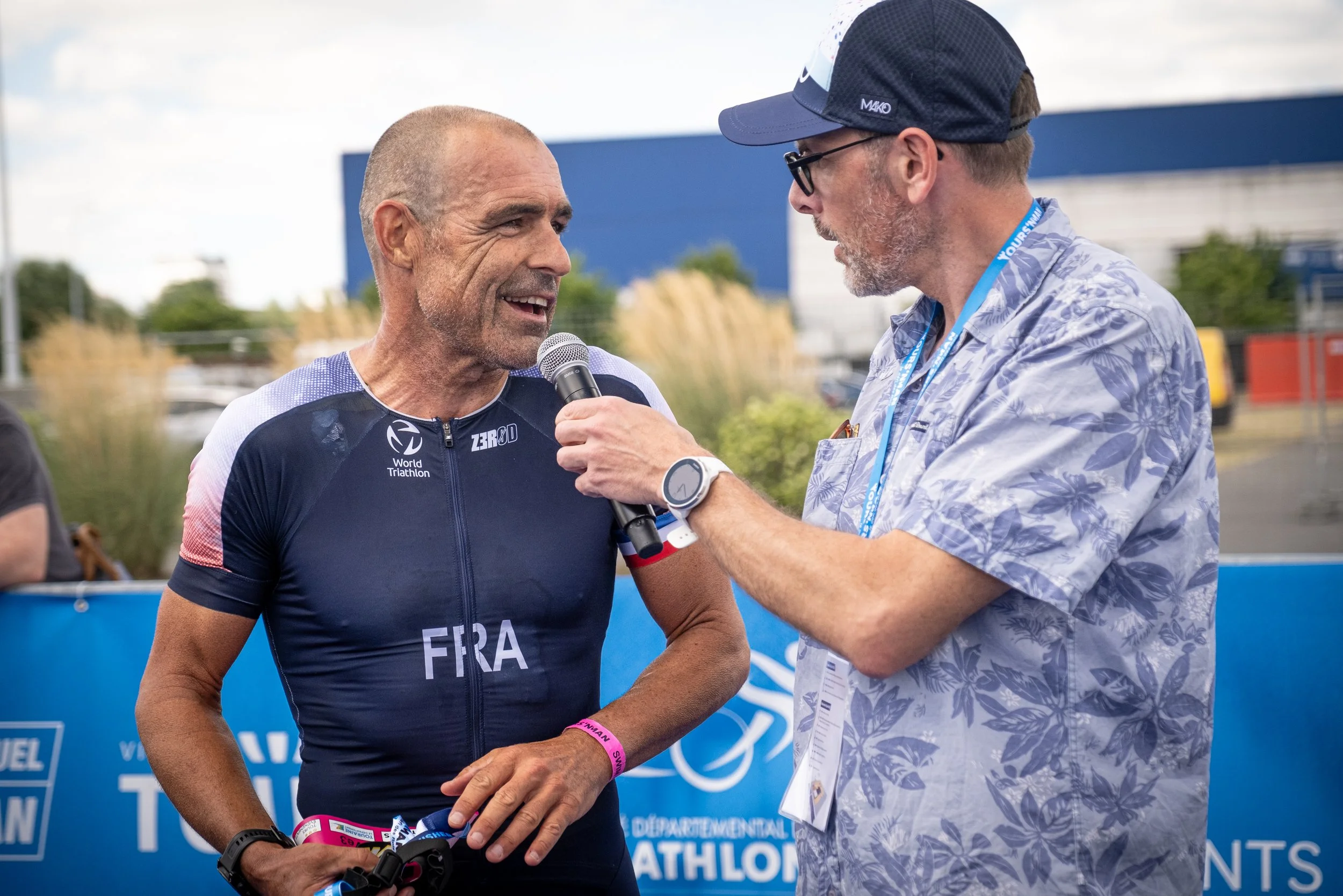 Un homme en tenue de triathlon, portant un maillot avec la mention 'FRA', est interviewé par un autre homme portant un chapeau et des lunettes, lors d'un événement en plein air.