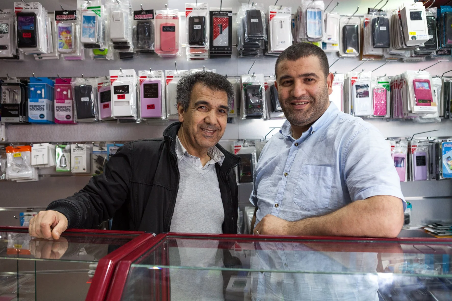 Deux hommes souriants dans une boutique de téléphones ou accessoires mobiles, debout derrière un comptoir en verre, avec un mur en arrière-plan rempli d'étuis et d'accessoires pour téléphones mobiles.