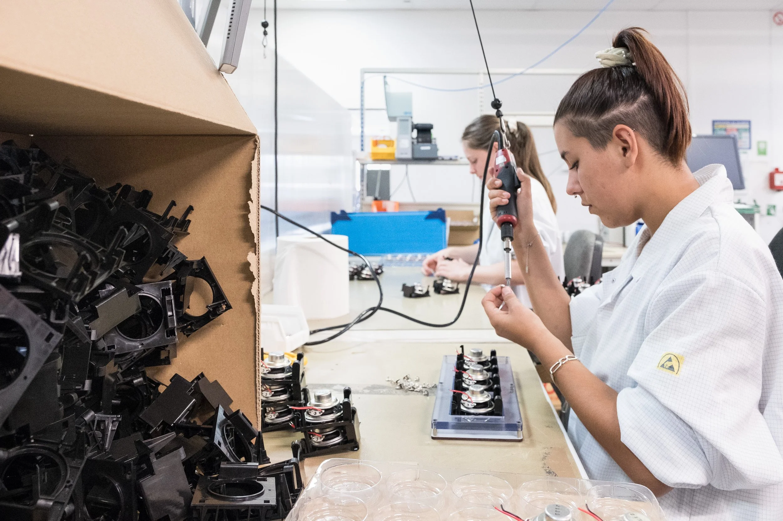Deux femmes travaillent sur une chaine de production, en utilisant un tournevis électrique pour assembler des composants électroniques.
