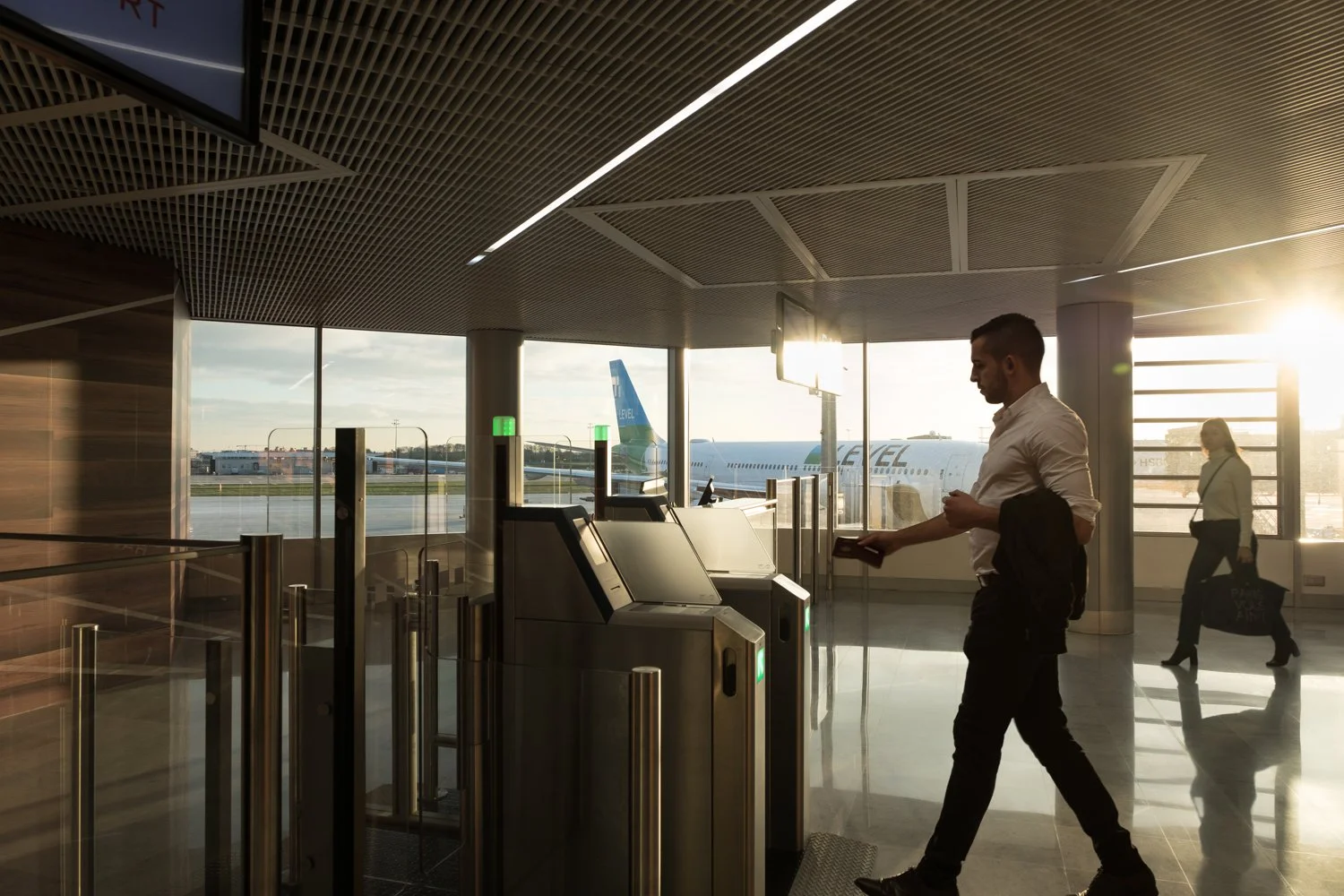 Un homme avec une chemise blanche et un pantalon noir se tient devant une borne d'enregistrement à l'aéroport, avec un avion en arrière-plan et le soleil couchant qui brille à travers les fenêtres.