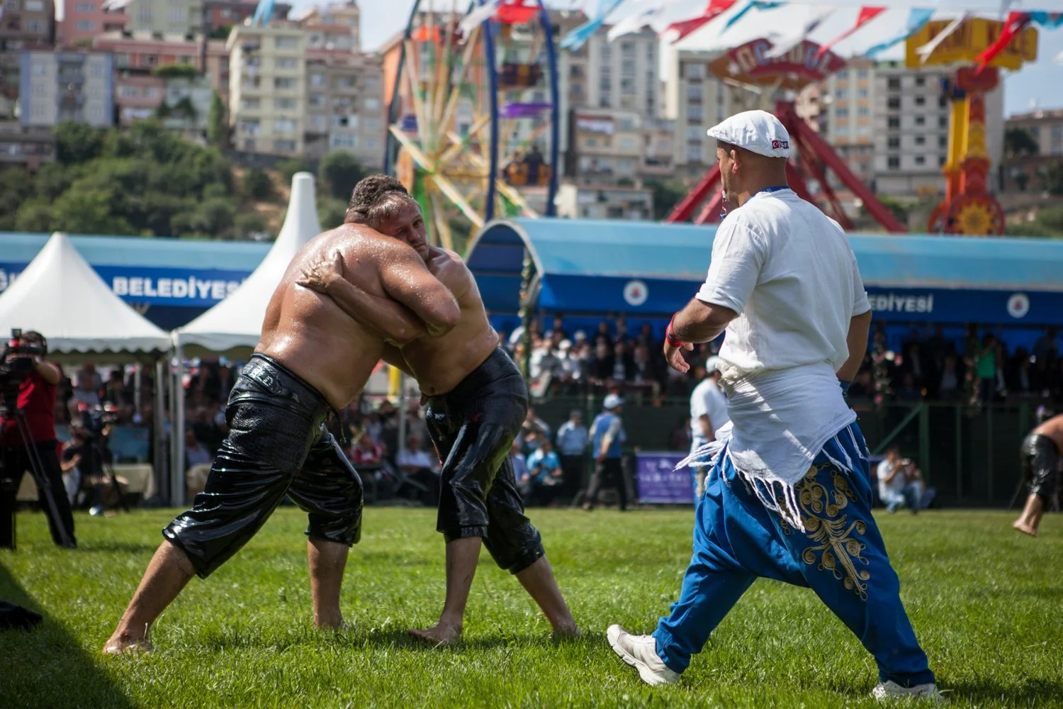 Deux hommes torse nus et huileux, en lutte. Un autre homme, habillé en blanc et bleu, arbitre sur le côté. En arrière-plan, des tentes blanches et une foule de spectateurs.