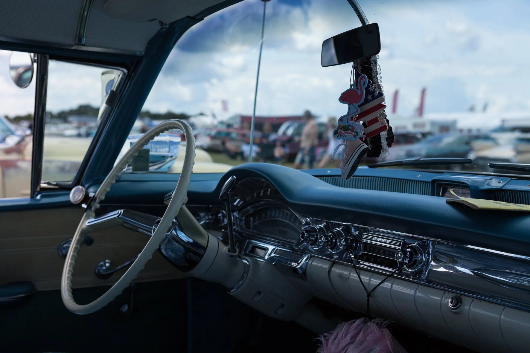 L'intérieur d'une voiture vintage avec un tableau de bord chromé, un volant blanc, des décorations suspendues au miroir, et des voitures en arrière-plan dans un parking extérieur.