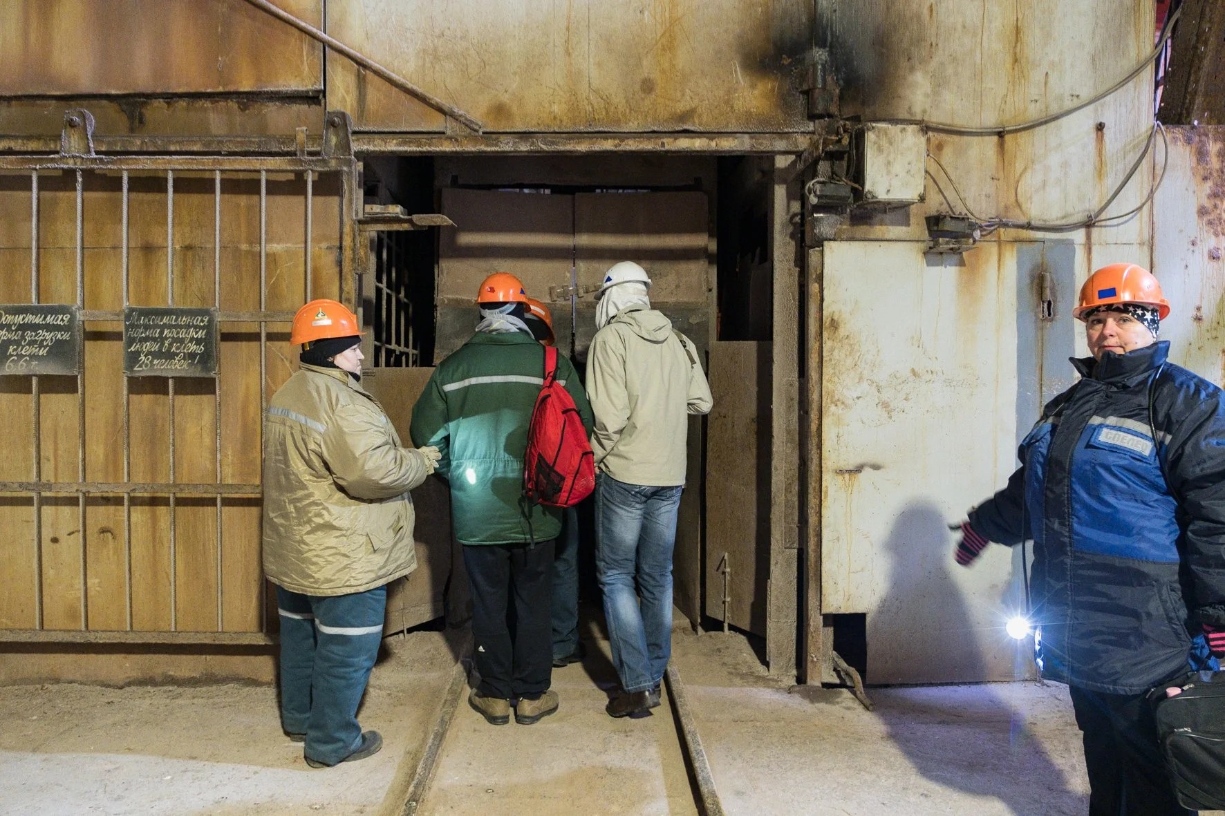 Groupe de personnes portant des casques orange et des vêtements de protection, entrant dans une mine ou un tunnel souterrain, avec une femme portant une veste noire et un casque bleu, tenant une lampe de poche.