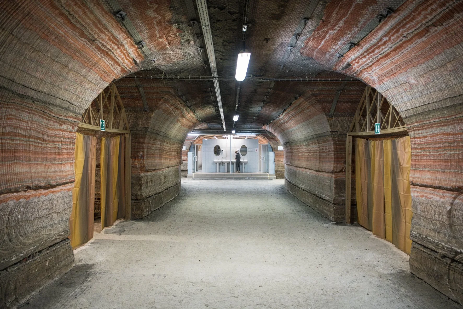 Un tunnel souterrain avec un plafond en roche rouge et deux entrées séparées par des rideaux de plastique jaune, menant à une zone avec des toilettes publiques en arrière-plan, éclairée par des lampes fluorescent.