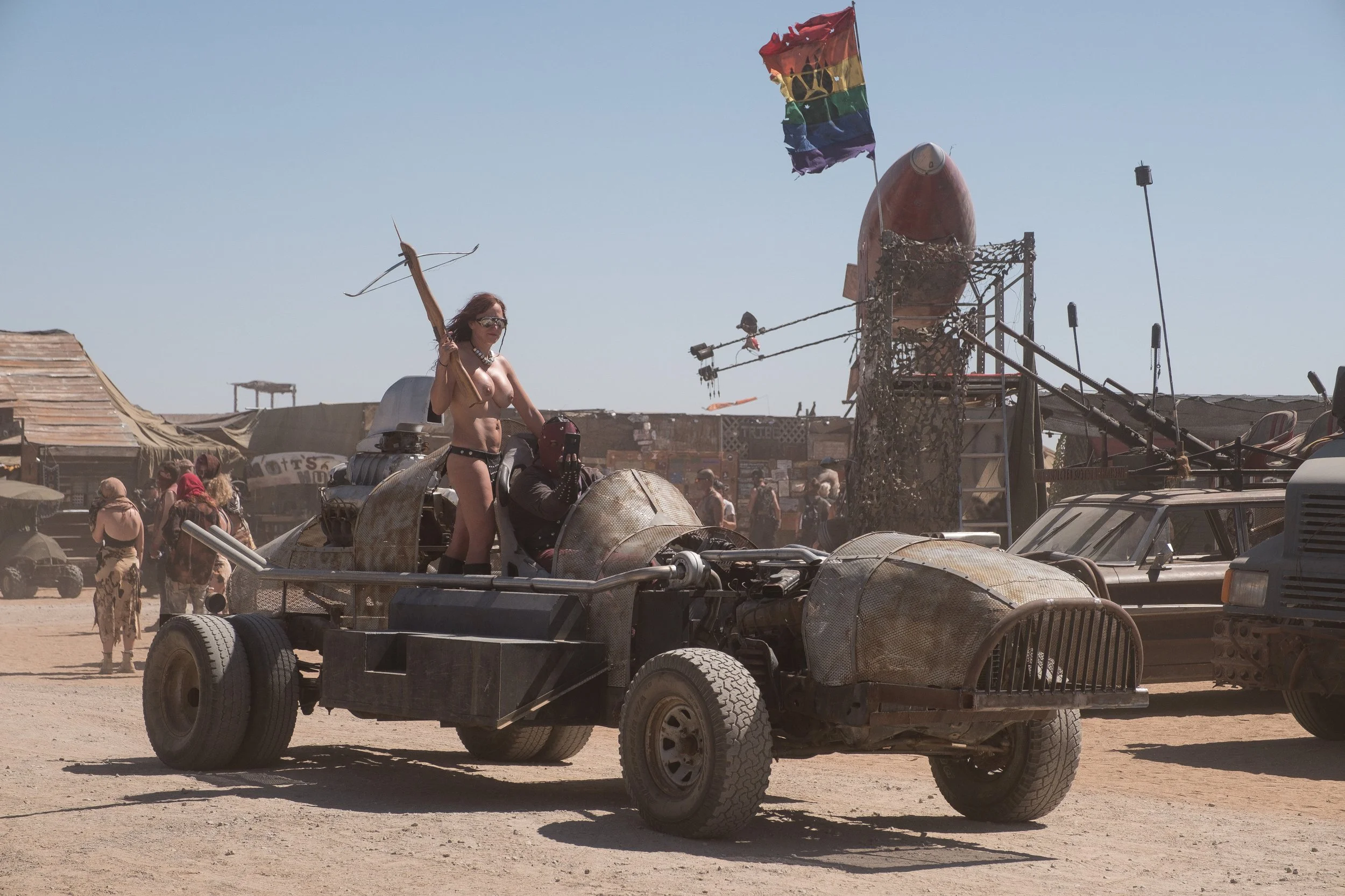Une femme nue portant des lunettes de soleil, tenant un arc, se tient sur un véhicule de style buggy dans un désert. En arrière-plan, des structures en bois et des personnes déguisées évoquant un univers post-apocalyptique au festival Wasteland.