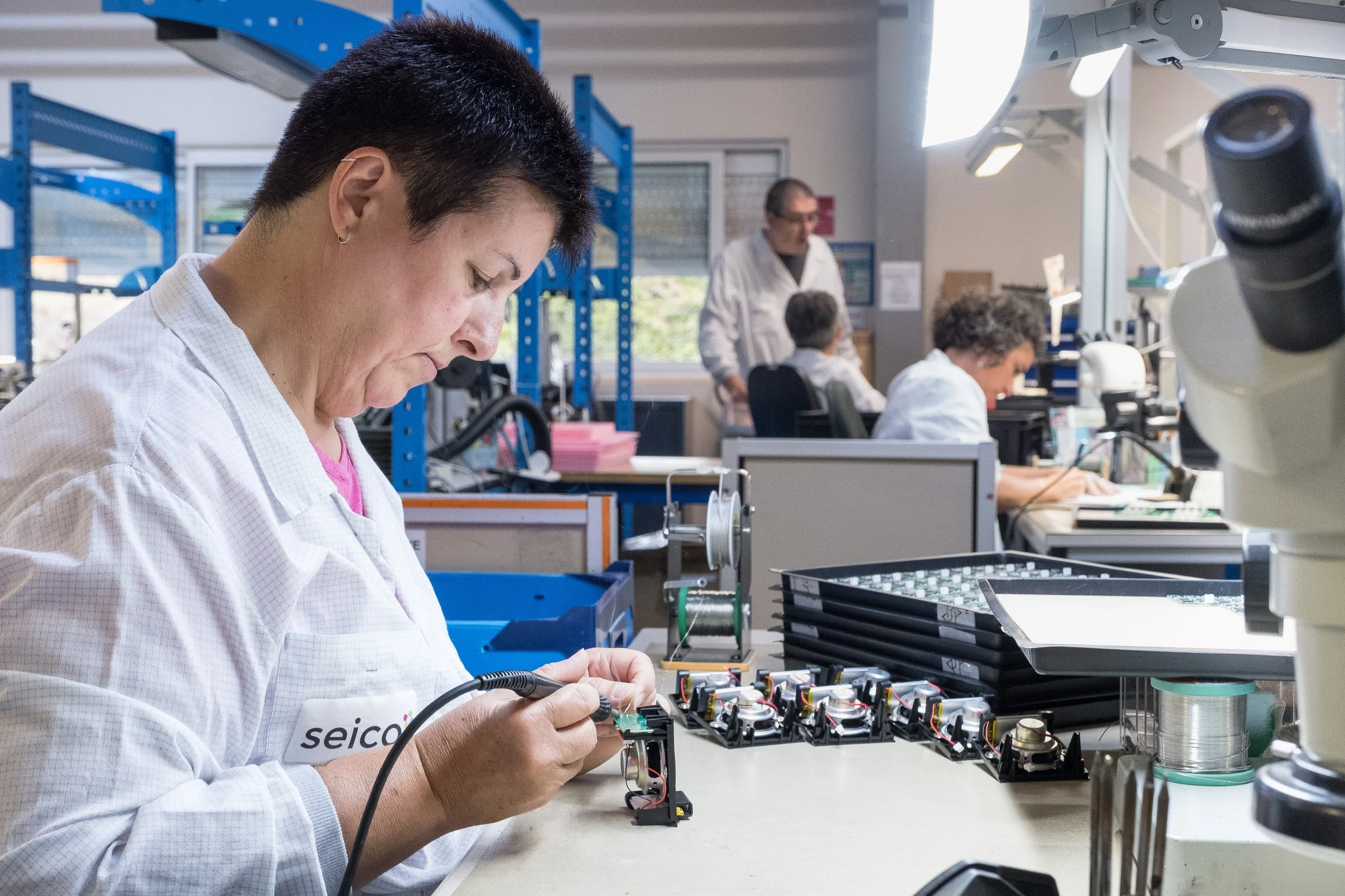 Une technicienne sur une chaine de production, portant un uniforme blanc, travaille sur un circuit électronique dans un environnement scientifique ou industriel avec d'autres travailleurs en arrière-plan.