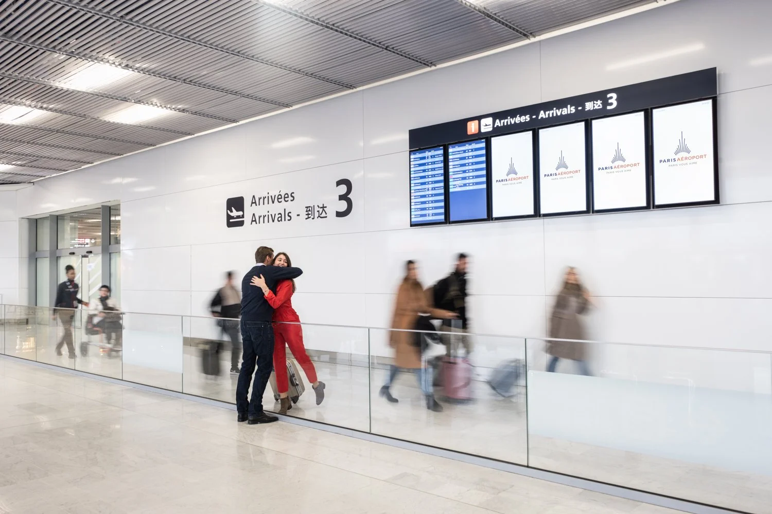 Deux personnes s'embrassent à l'aéroport devant un panneau indiquant les zones d'arrivée, avec d'autres passagers passant en arrière-plan.