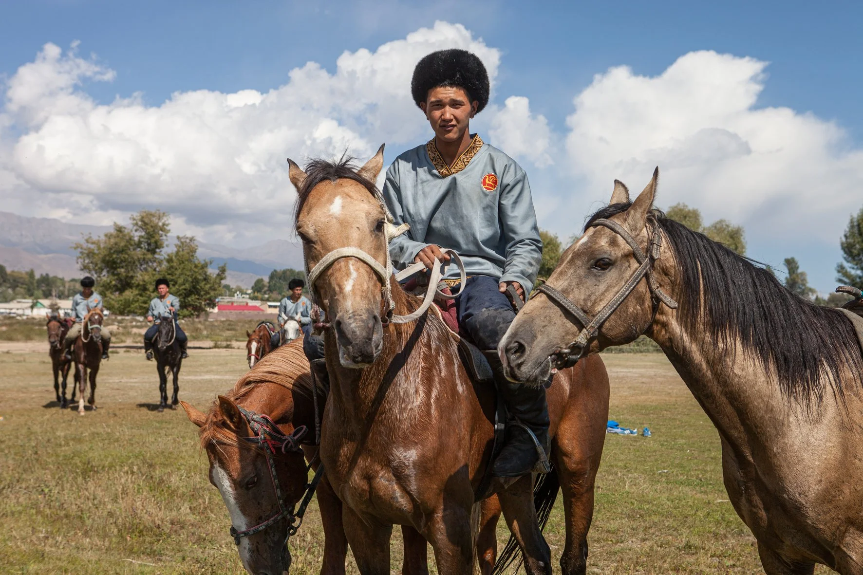 Jeune homme asiatique en costume traditionnel mongol à cheval, entouré d'autres cavaliers, dans un paysage de plaines avec montagnes en arrière-plan.