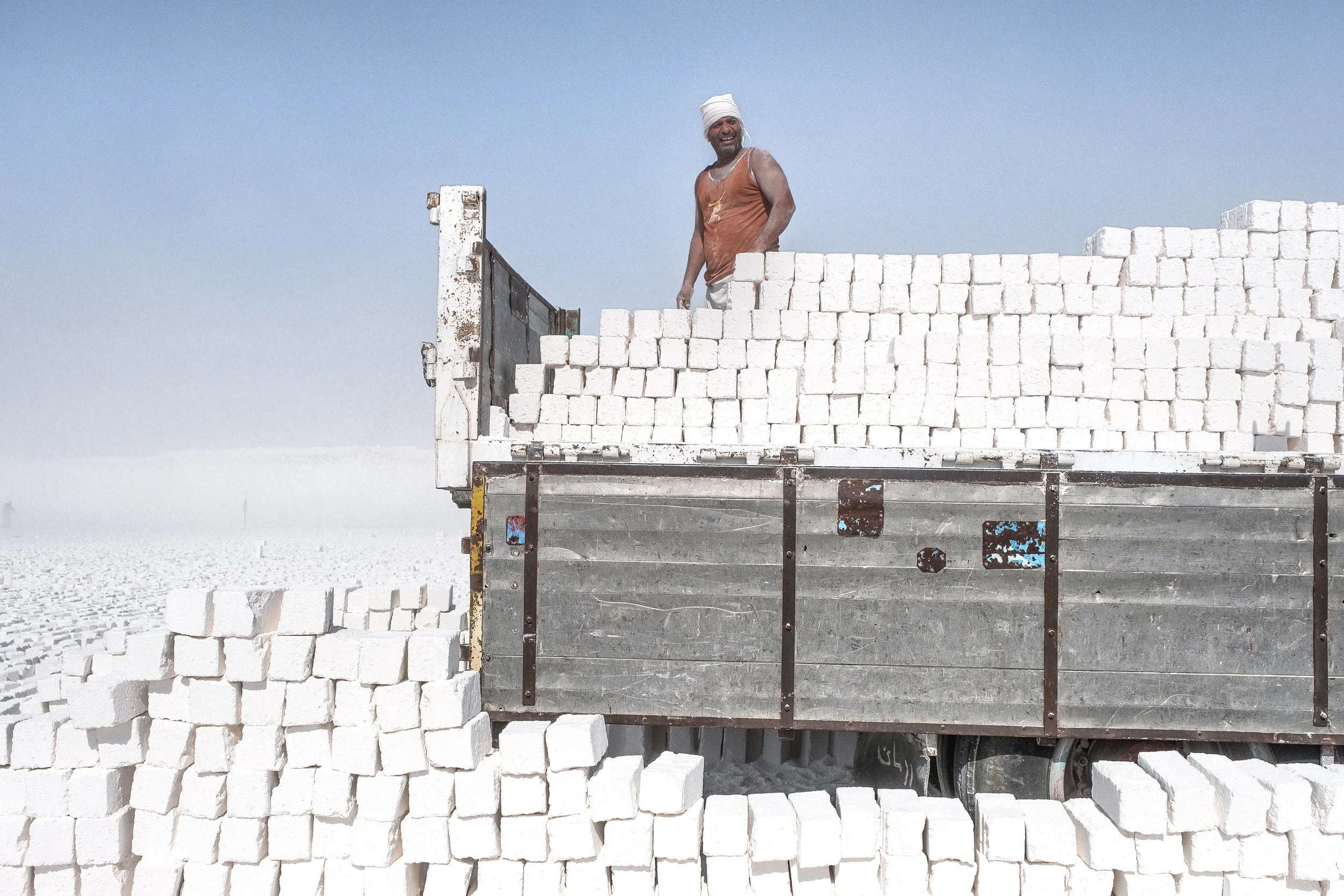 Homme souriant portant un turban blanc, debout dans une ferme de blocs de calcaire blancs, sous un ciel clair.