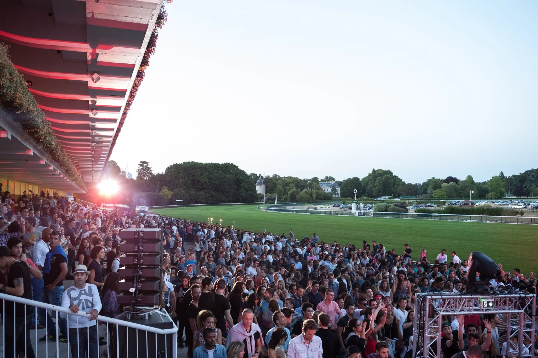 Une foule de spectateurs assiste à un événement en plein air à l'hippodrome de Longchamp, avec une pelouse verte et des arbres en arrière-plan, lors du coucher du soleil.
