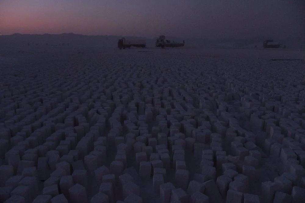 Paysage désertique avec des formations rocheuses de couleur claire sous un ciel sombre au crépuscule, avec des véhicules ou structures dispersés à l'horizon.