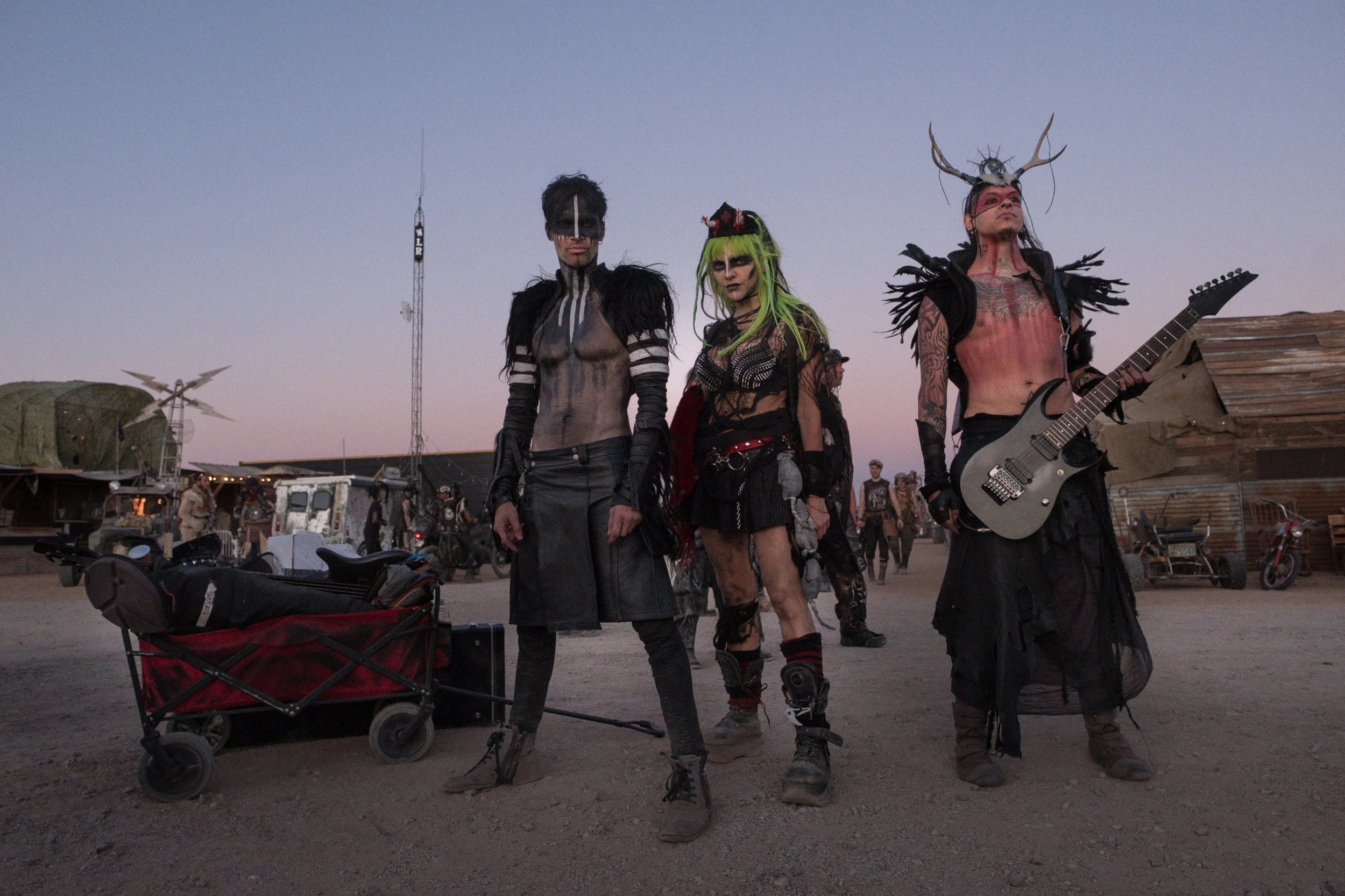 Trois personnes en costumes de festival futuristes et gothiques avec maquillage élaboré dans un désert, avec des véhicules en arrière-plan.