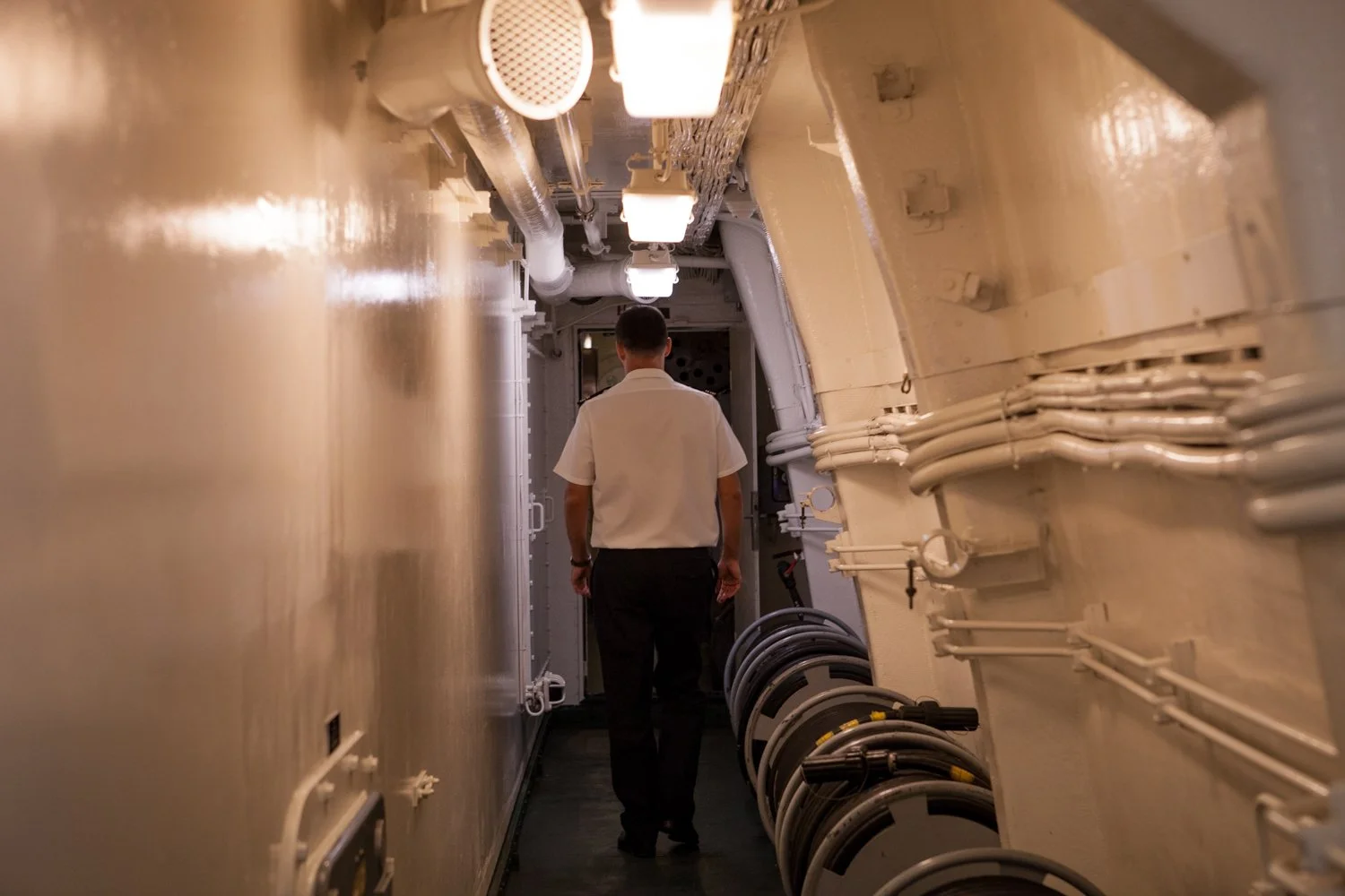 Le commandant en uniforme blanc marche dans un couloir étroit, à l'intérieur de la frégate militaire Surcouf, avec des tuyaux métalliques et des câbles visibles au plafond et sur les murs.