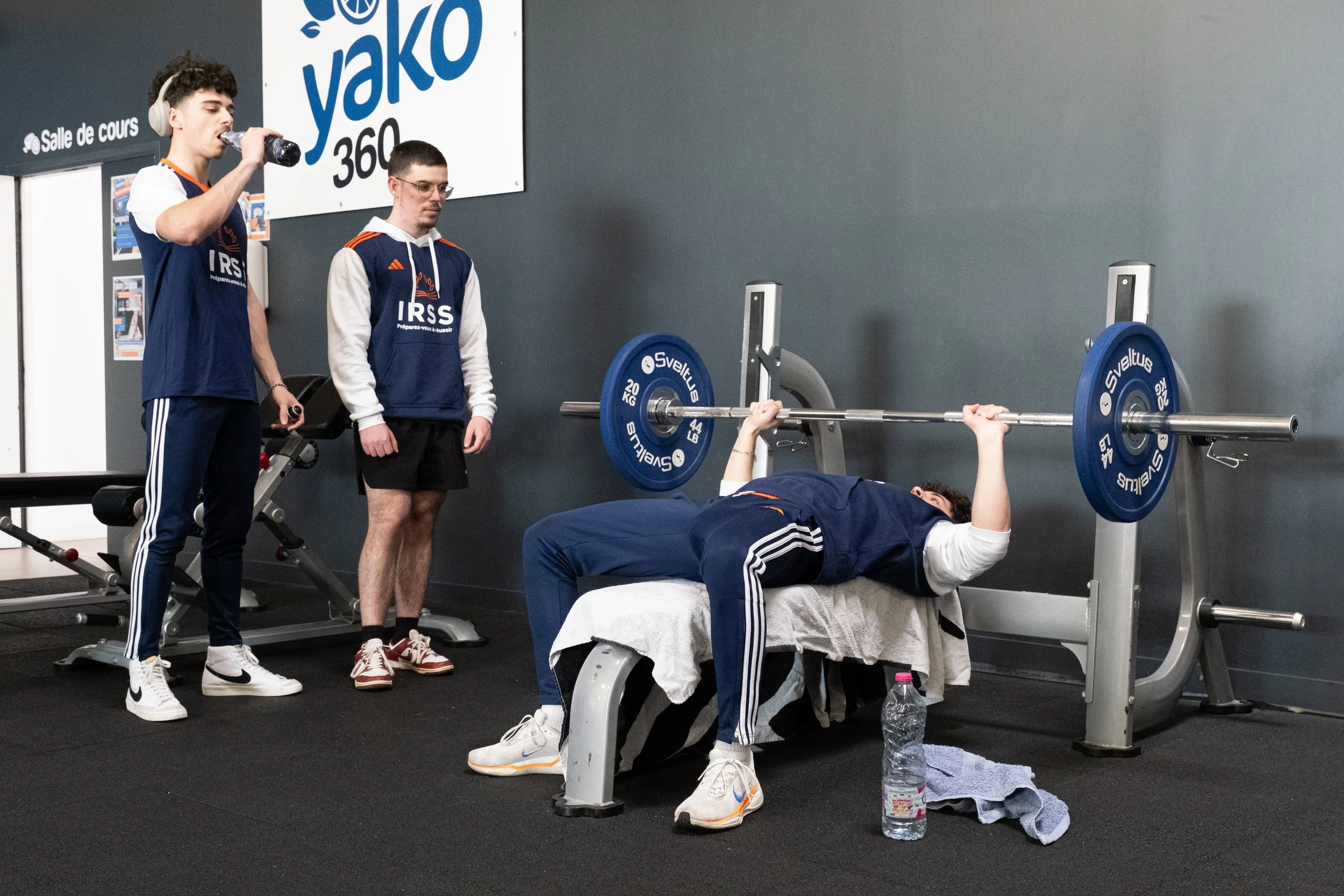Un homme en tenue de sport soulève une barre chargée de poids lors d'une séance d'entraînement, entouré de deux autres hommes, l'un buvant de l'eau et l'autre regardant, dans une salle de sport.