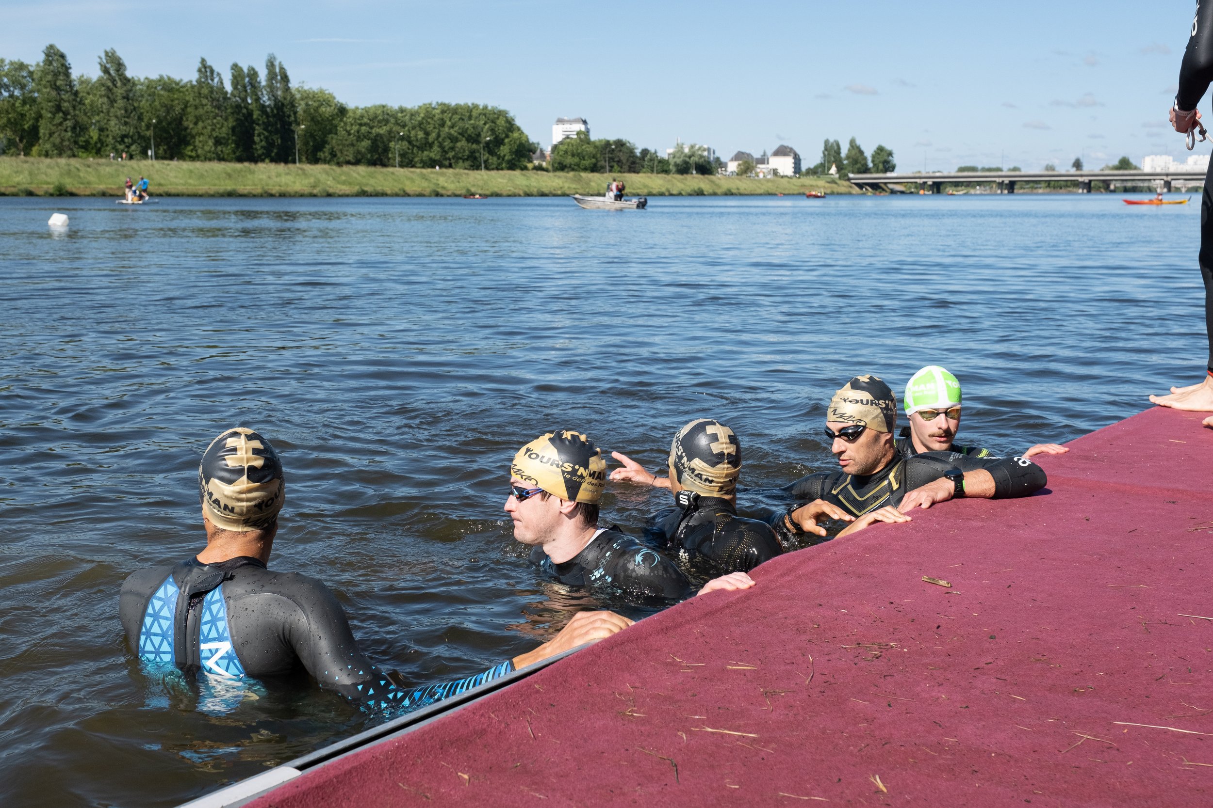 Groupe de nageurs en train de sortir de l'eau à une station de triathlon dans un lac ou une rivière, portant des combinaisons de natation et des bonnets.