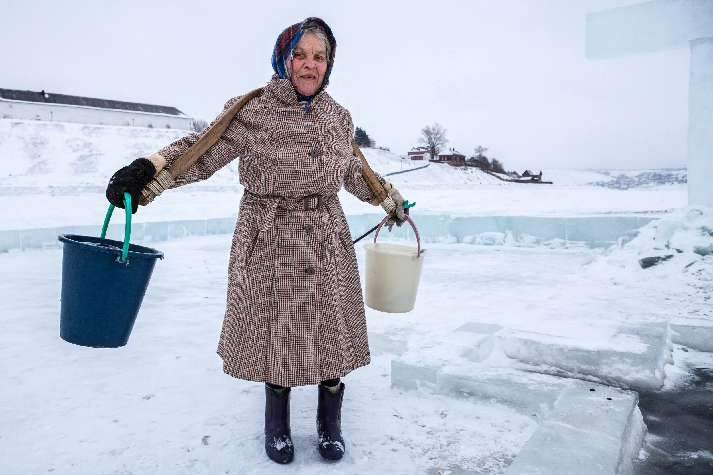 Femme âgée portant un manteau à carreaux, un foulard en laine, des gants et des bottes en caoutchouc, tenant des seaux dans un environnement hivernal avec de la glace et de la neige.