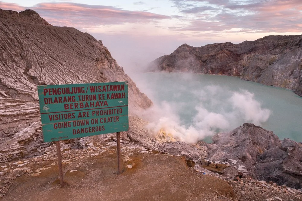 Vue du cratère volcanique du Kawah Ijen avec de la vapeur s'échappant, un panneau d'avertissement en indonésien et en anglais, indiquant que la zone est dangereuse et interdite aux visiteurs.