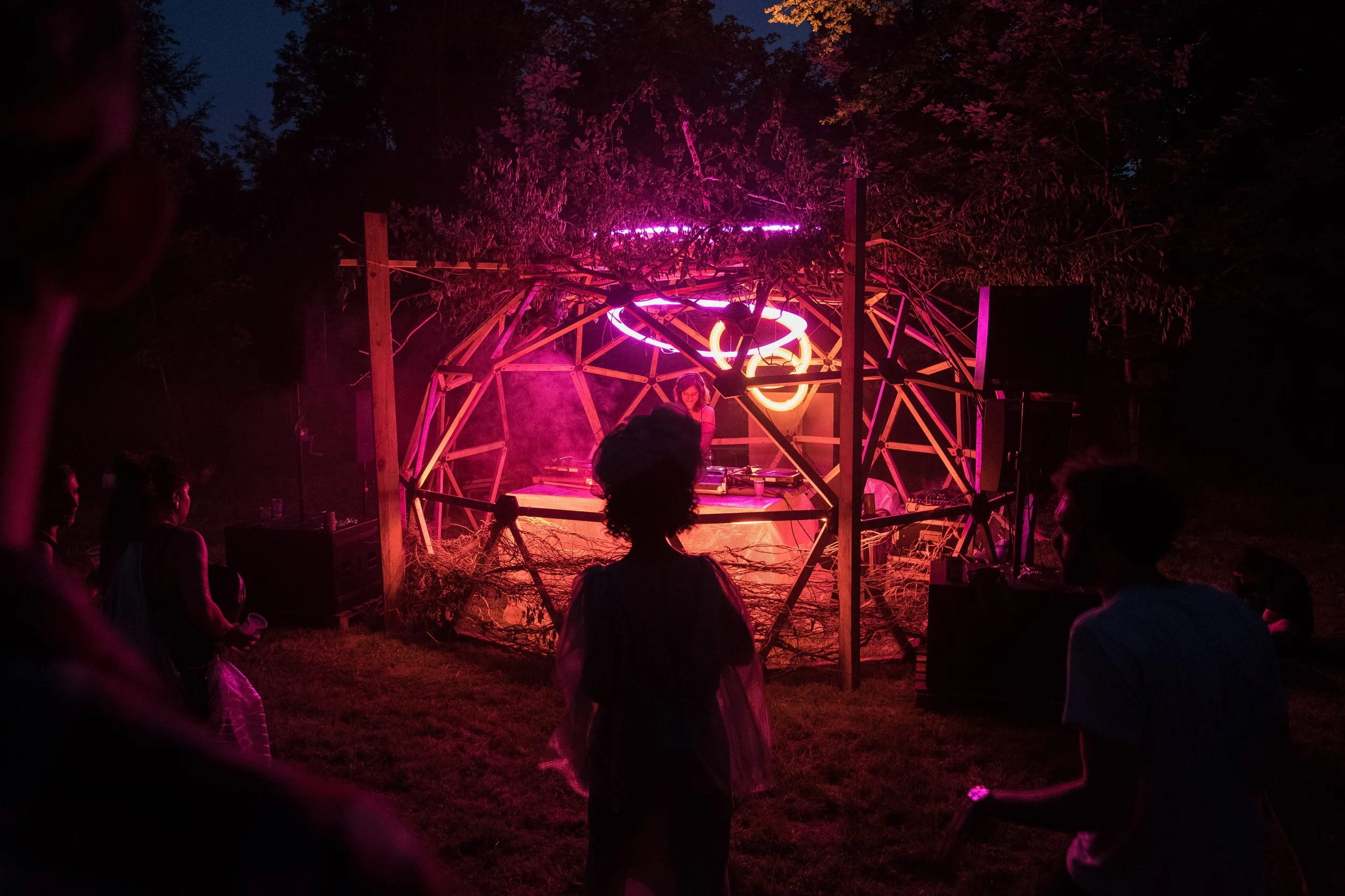 Une scène de nuit avec un DJ jouant dans une structure géodésique éclairée de lumières roses et oranges, entourée de personnes qui dansent, dans un environnement extérieur avec des arbres en arrière-plan.