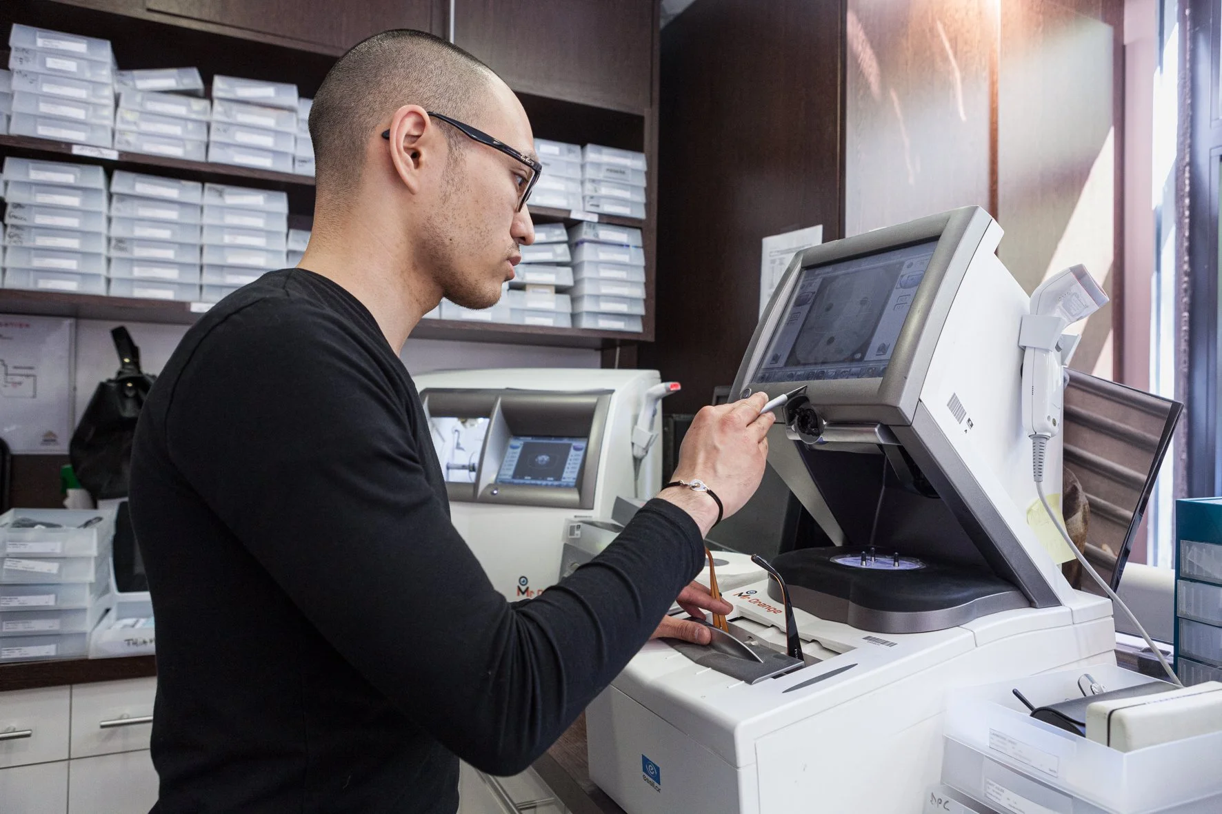 Un homme travaille dans un laboratoire ou une officine, utilisant un appareil de diagnostic médical devant un ordinateur. Il porte des lunettes et une couleur noire. Le fond montre des étagères avec des boîtes de rangement.