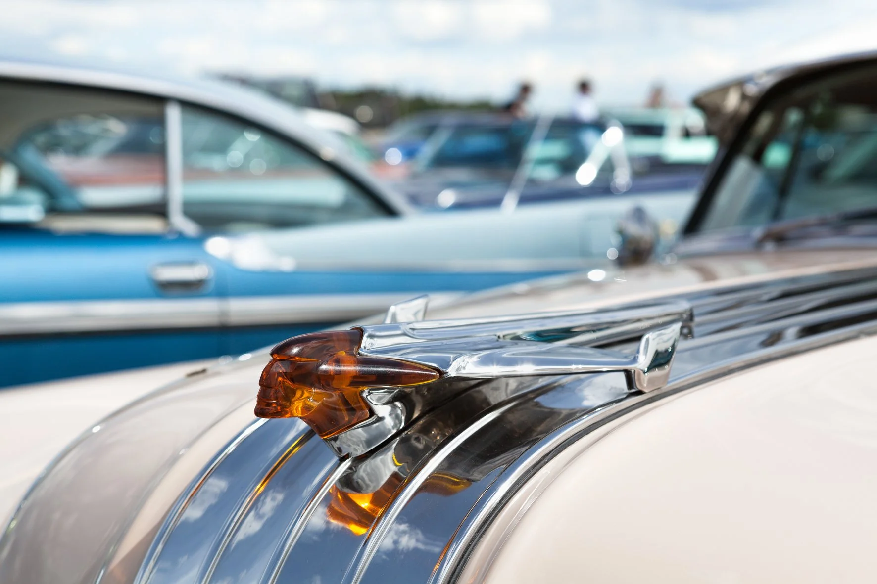 Gros plan d'une pièce décorative en forme de tête de lion en métal sur le capot d'une voiture classique, avec d'autres voitures vintage floues en arrière-plan.