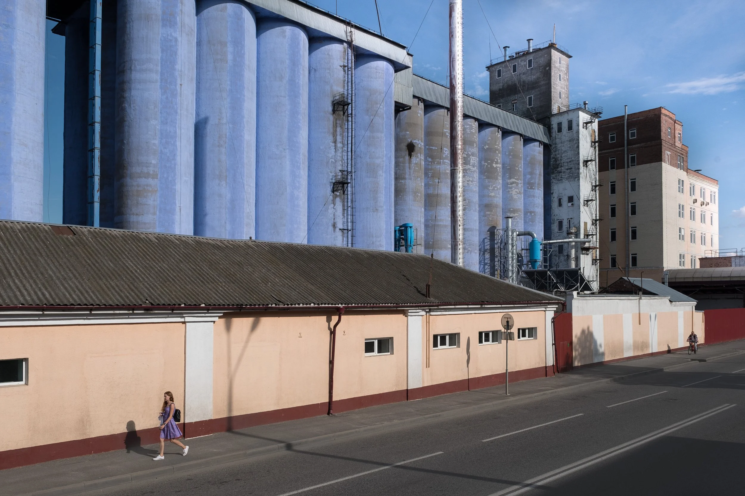 Une jeune femme marche le long d'un trottoir devant un bâtiment avec des silos industriels en arrière-plan, sous un ciel bleu avec quelques nuages en Biélorussie.