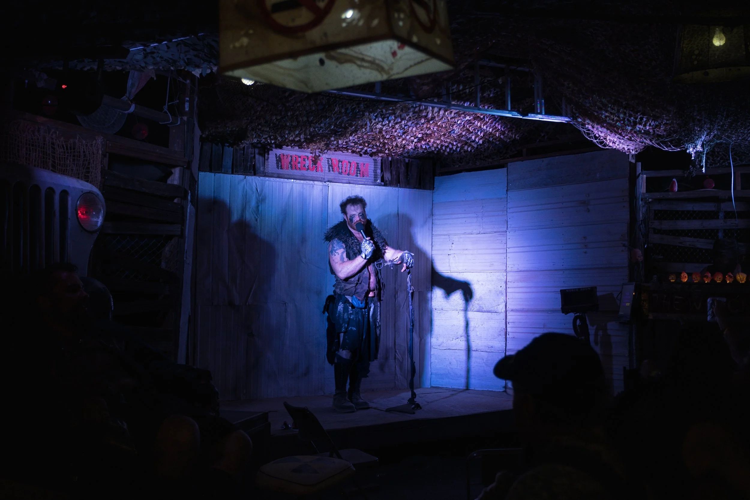 Un artiste chante lors d'un karaoké post-apocalyptique dans une pièce appelée 'Wreck Room', avec un décor rustique en bois, lumière violette et quelques spectateurs en silhouette dans l'obscurité.